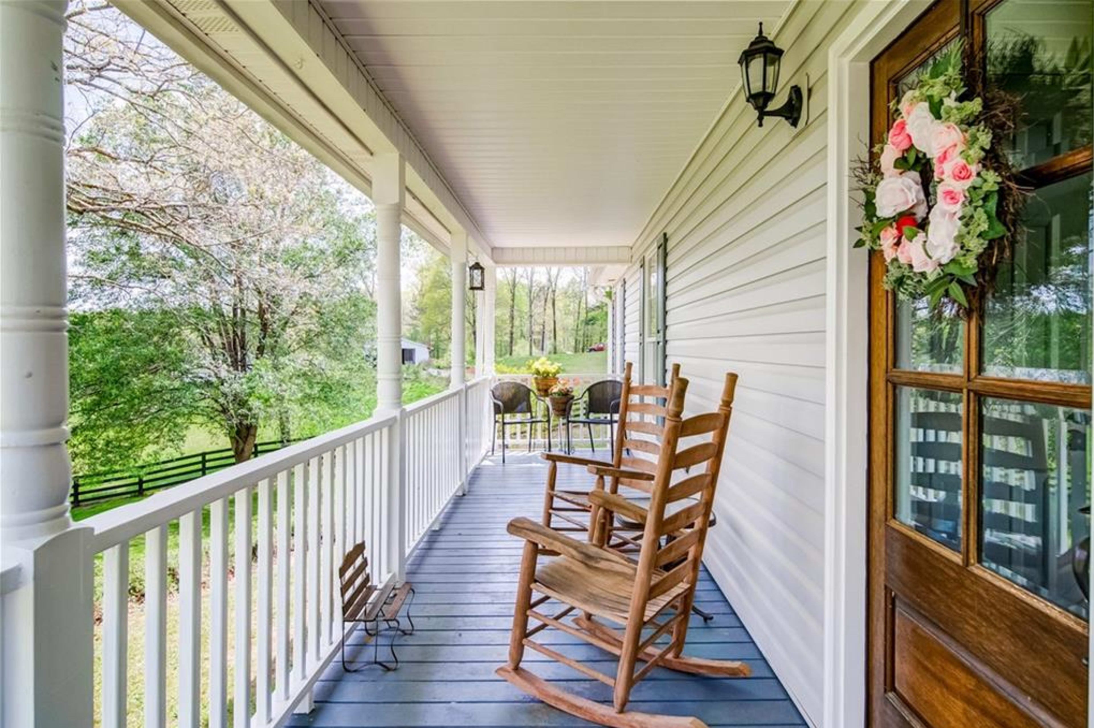 A porch with two rocking chairs and a small child's chair, surrounded by greenery and a view of trees in the background.