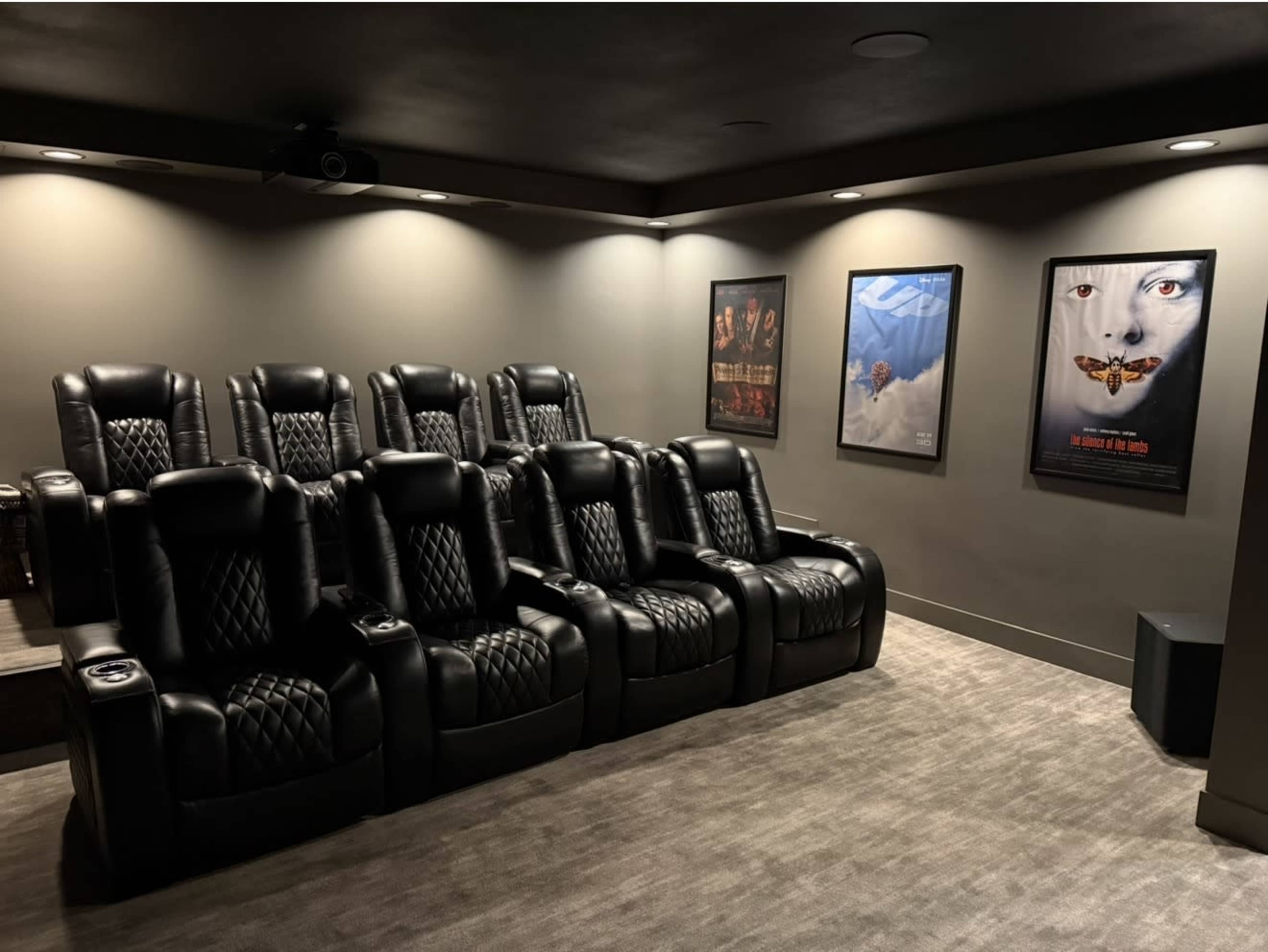 The image shows a home theater with six black leather reclining chairs arranged in front of wall-mounted movie posters and a projector.