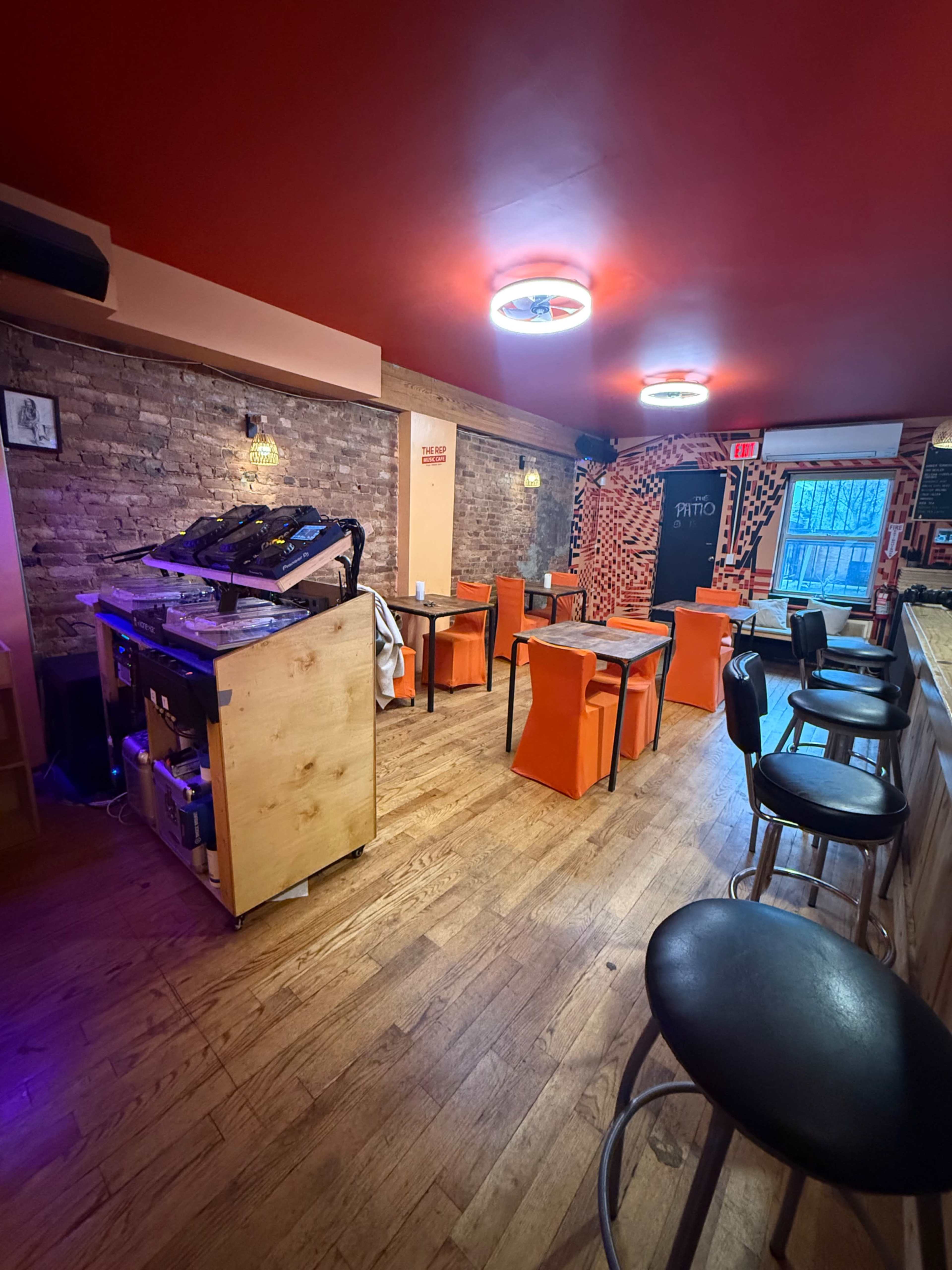 The image shows a restaurant interior with wooden flooring, orange and black chairs, and a bar area.