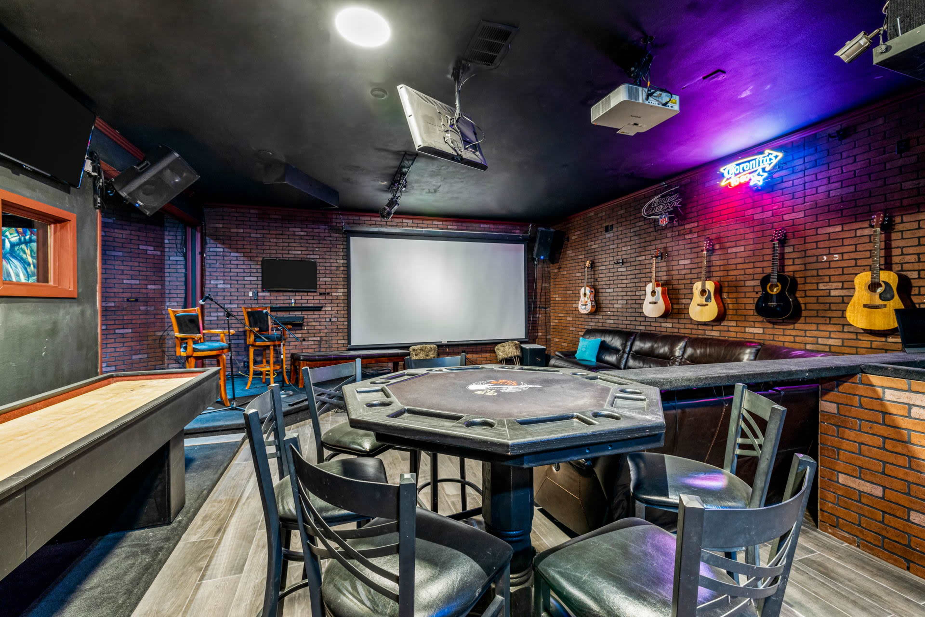 A game room features a poker table surrounded by chairs, a large projection screen, and wall-mounted guitars, all set against a brick backdrop.