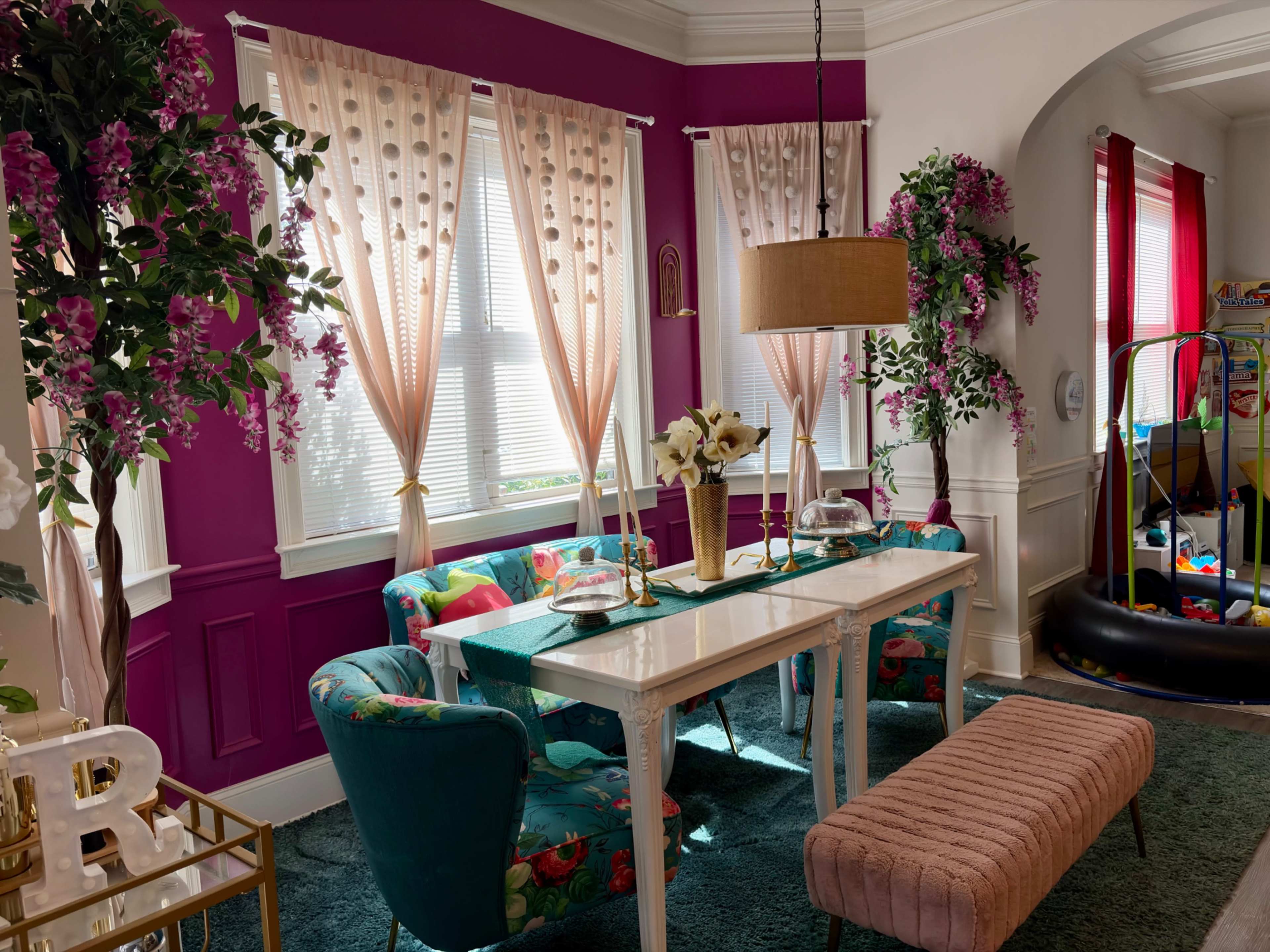 The image shows a brightly decorated living space with a white table, colorful chairs, potted plants, and large windows adorned with sheer curtains.