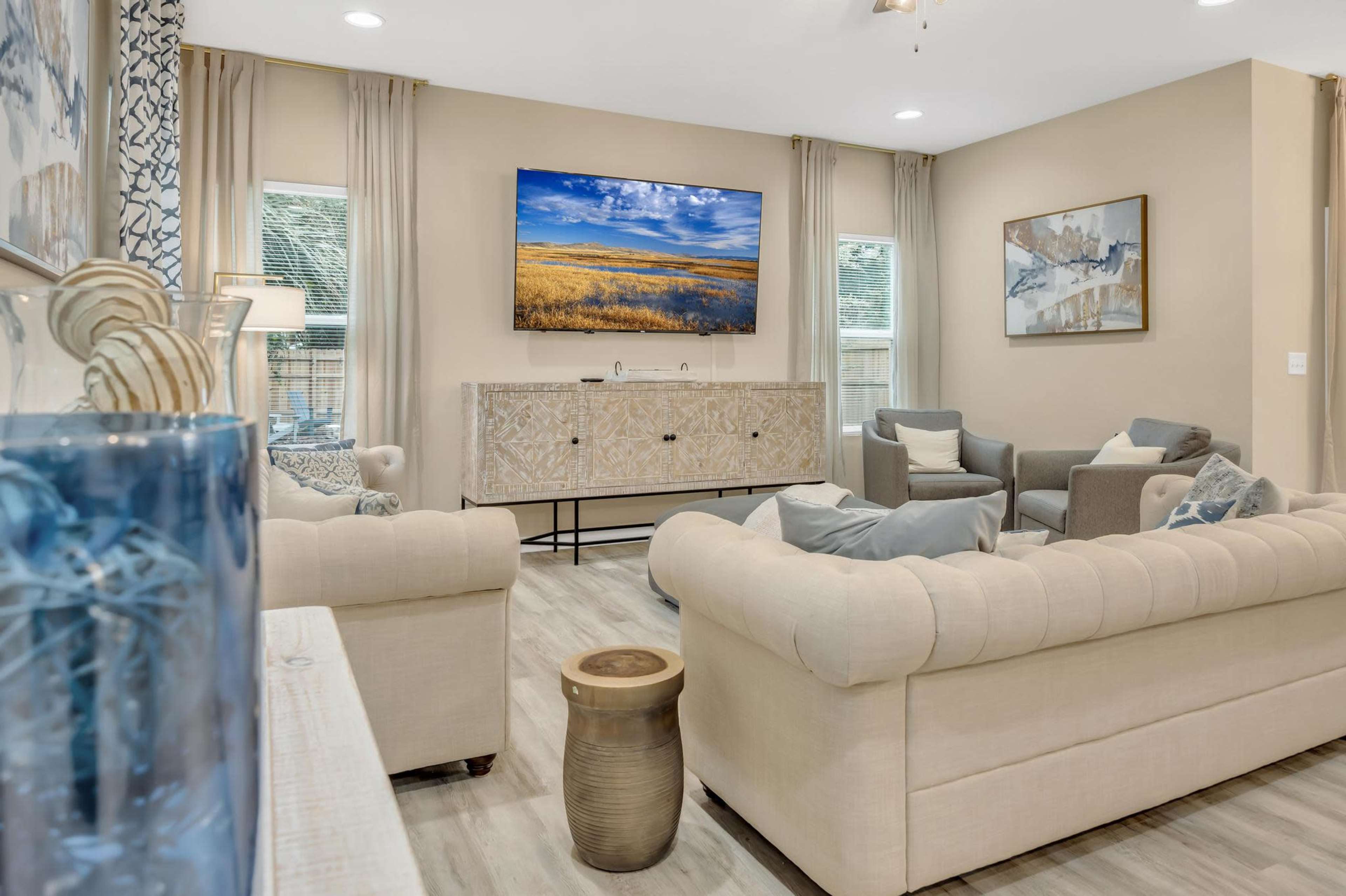 The living room features a light-colored sofa and armchairs arranged around a wooden coffee table, with a large wall-mounted TV displaying a landscape image.