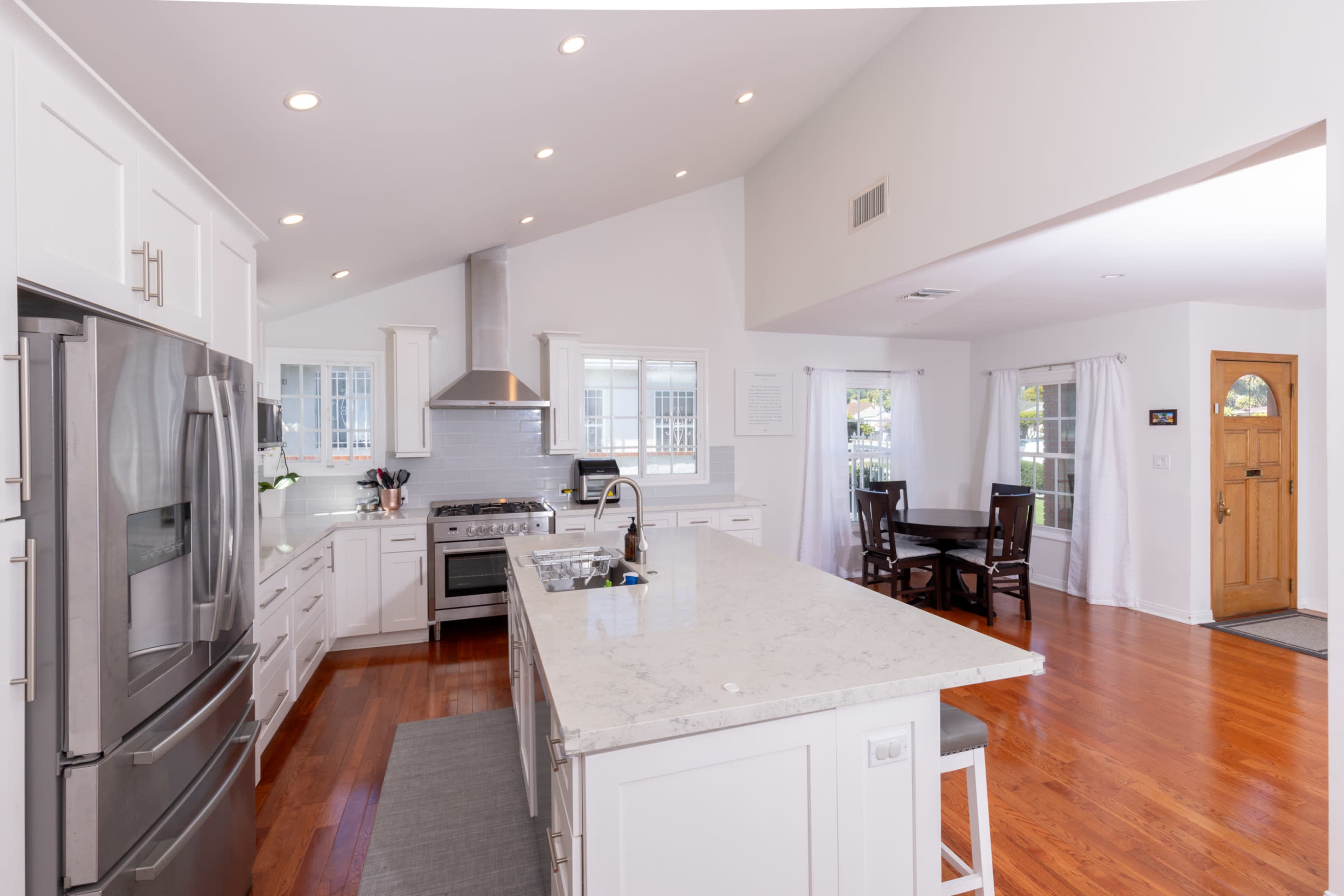 The image shows a modern kitchen with white cabinetry, stainless steel appliances, a large island, and an adjacent dining area with a wooden table and chairs.