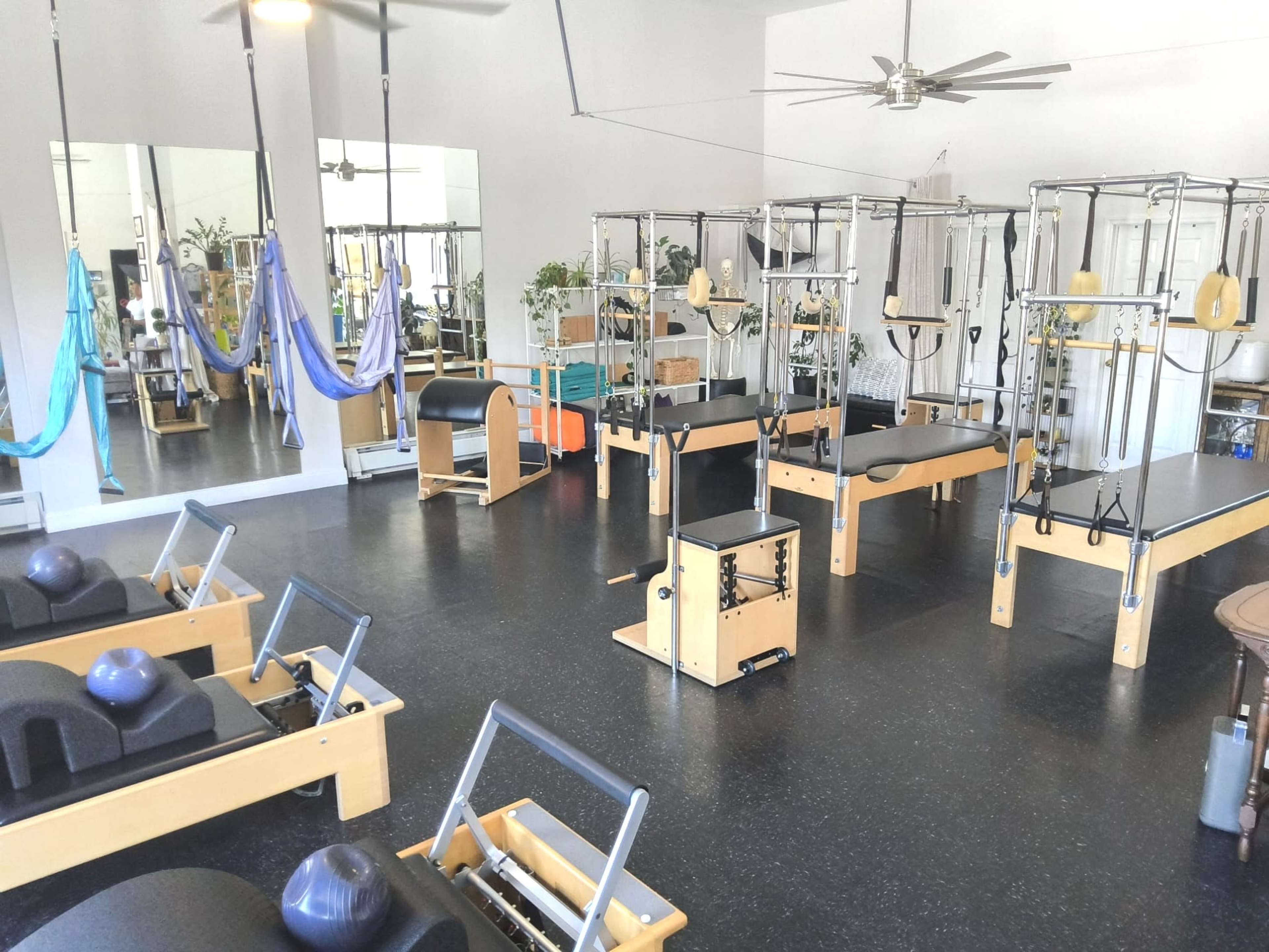 The image shows a spacious Pilates studio equipped with reformer machines, mats, and aerial yoga hammocks arranged neatly in the room.