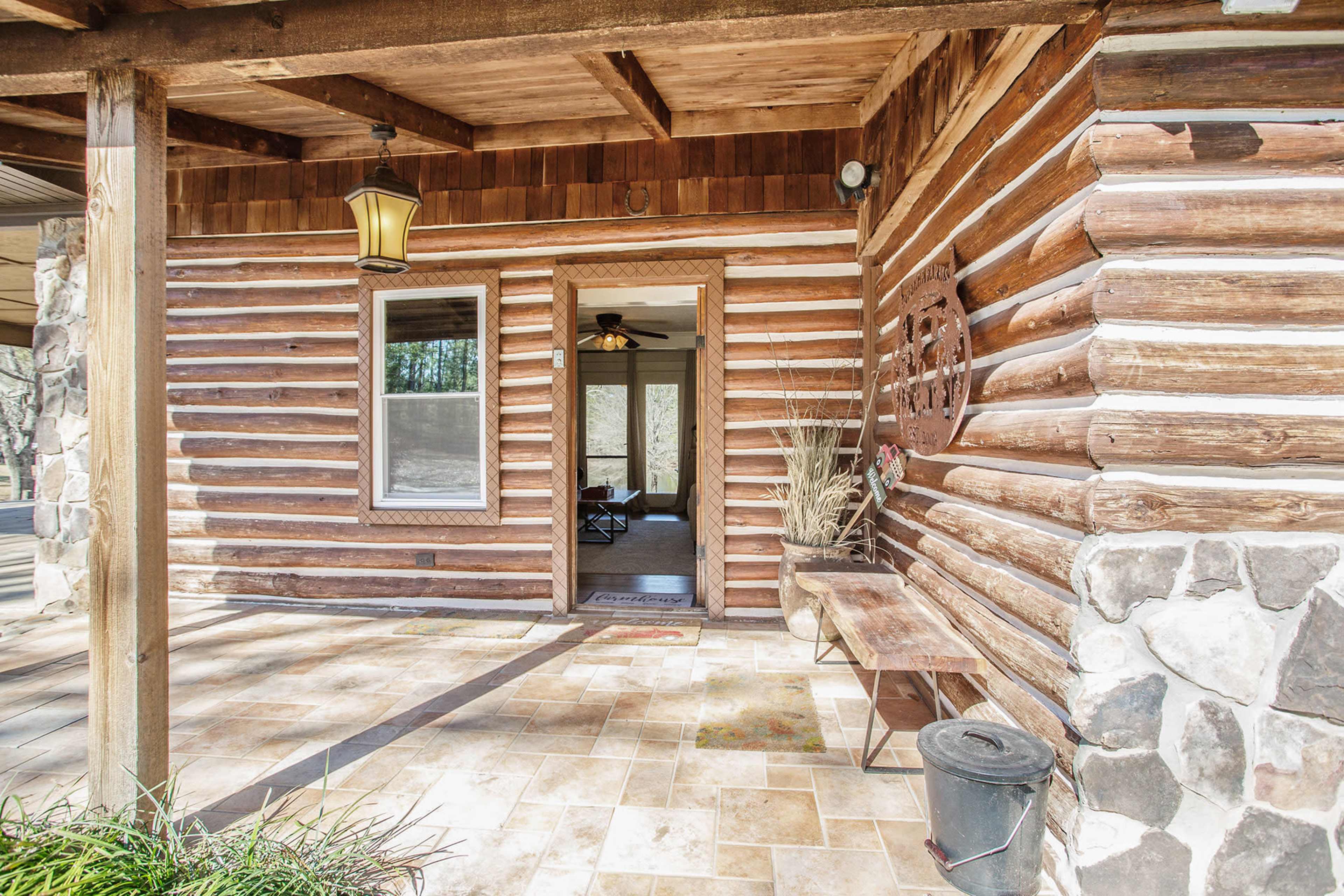 The image shows a wooden log cabin entrance with a stone patio, an open door leading into the interior, and a small table beside the entrance.