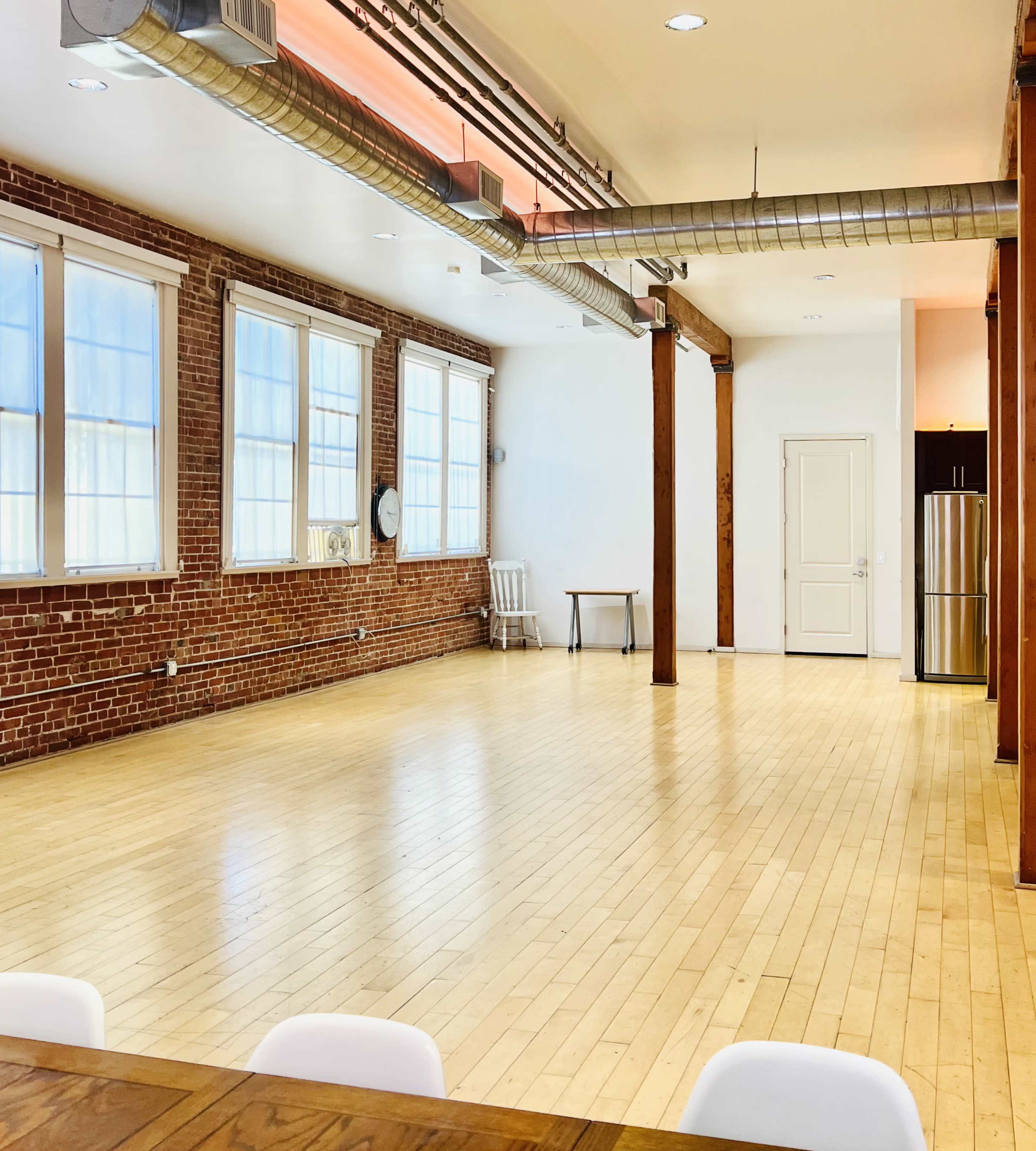 The image shows a spacious, modern room with exposed brick walls, wooden floors, and large windows, featuring minimal furniture including a table and a stainless steel refrigerator.