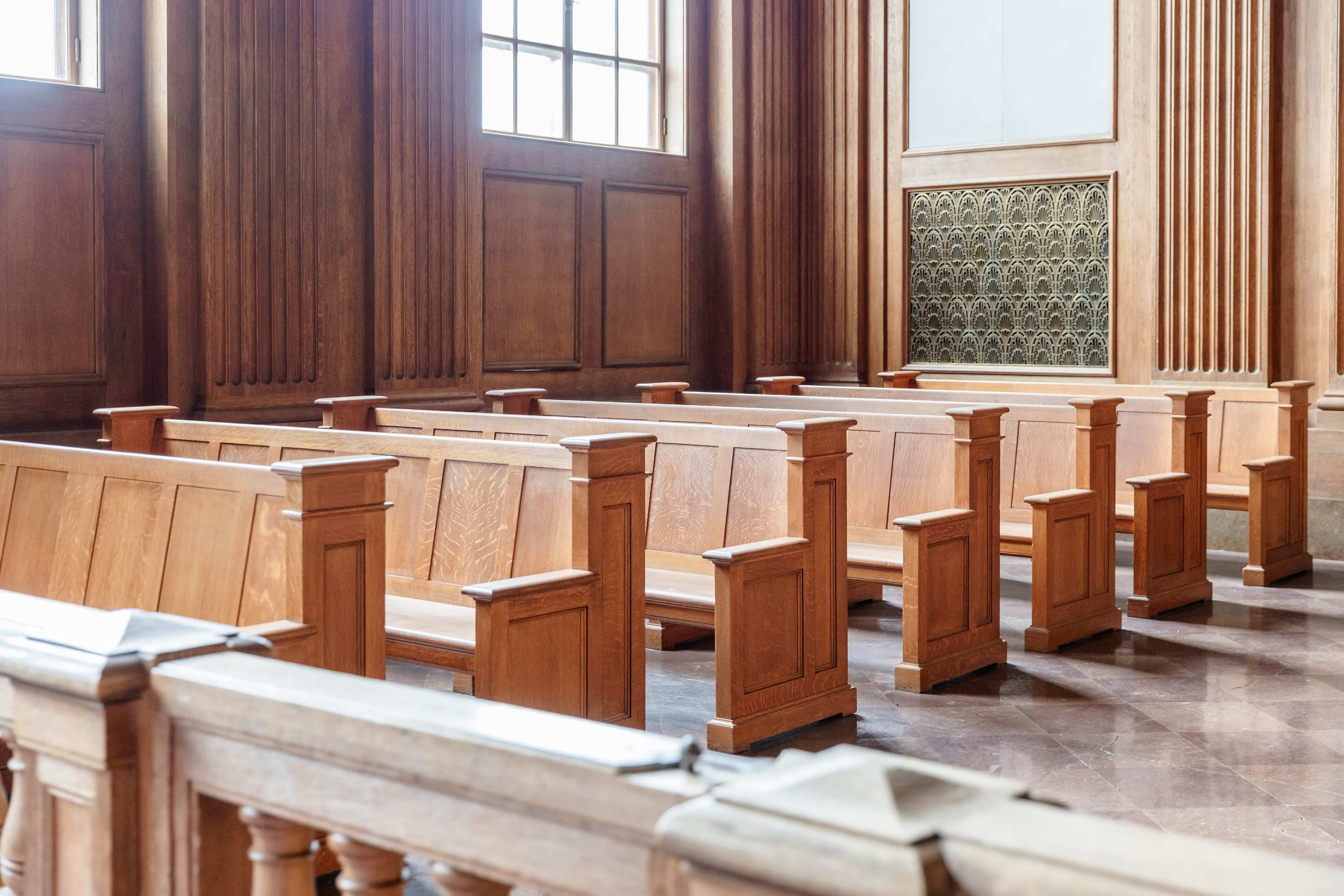 The image shows a wooden interior of a courtroom, featuring rows of wooden benches with a polished floor and large windows.