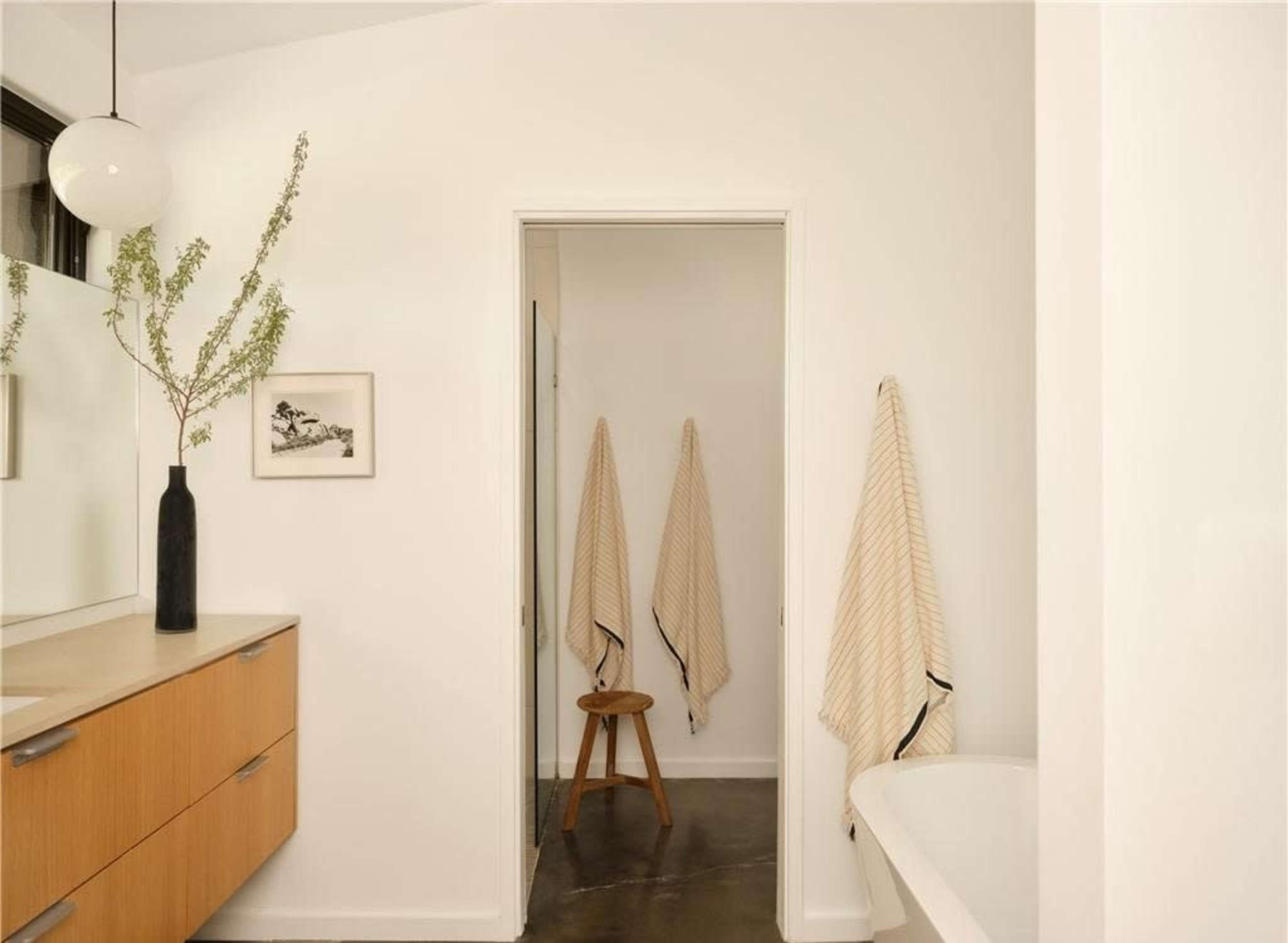 A modern bathroom features a wooden vanity, a potted plant, three hanging towels, and a stool next to a freestanding bathtub.