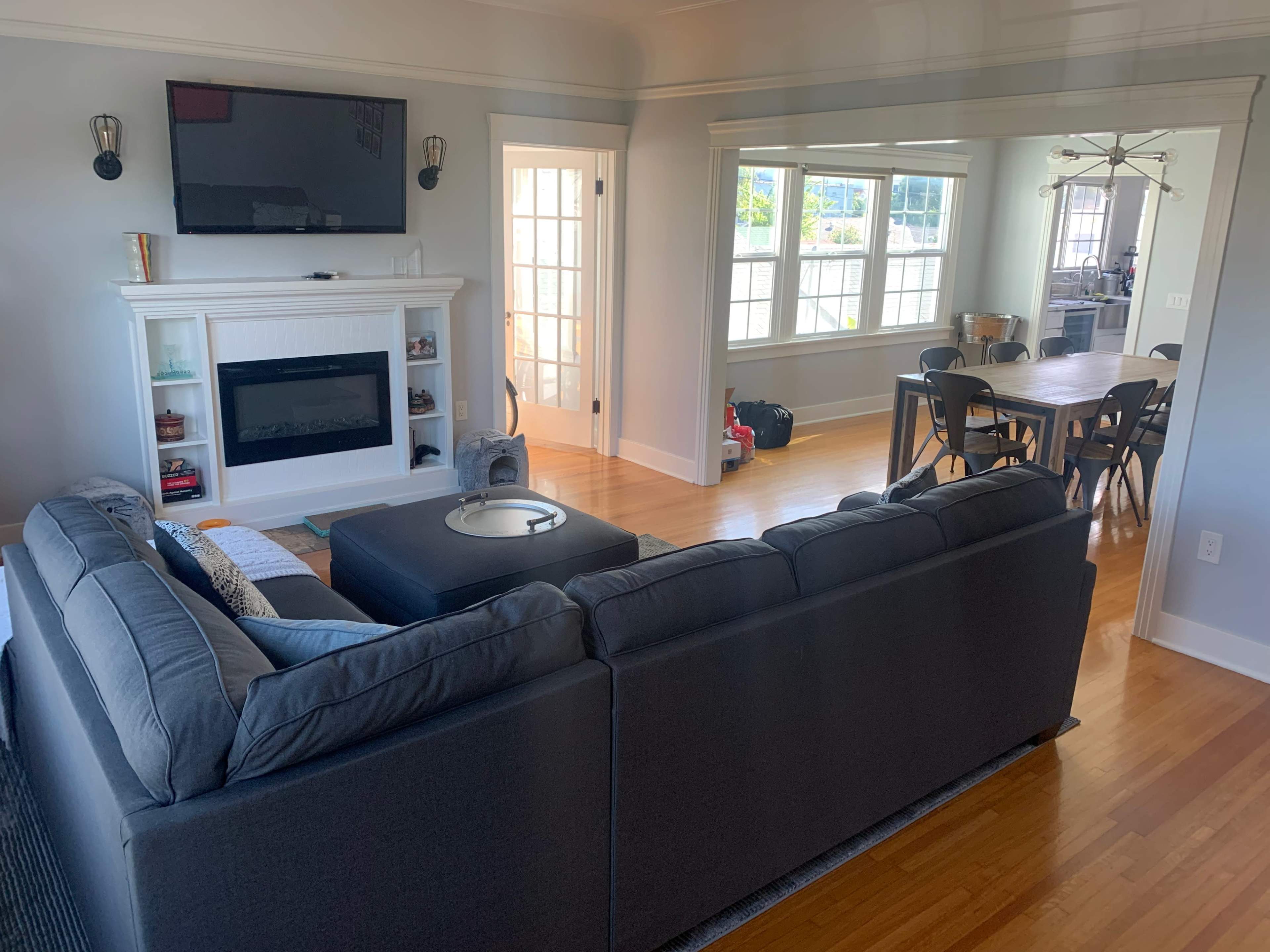 The image shows a living room with a gray sectional couch, a wall-mounted television above a white fireplace, and a dining area visible through an open doorway.