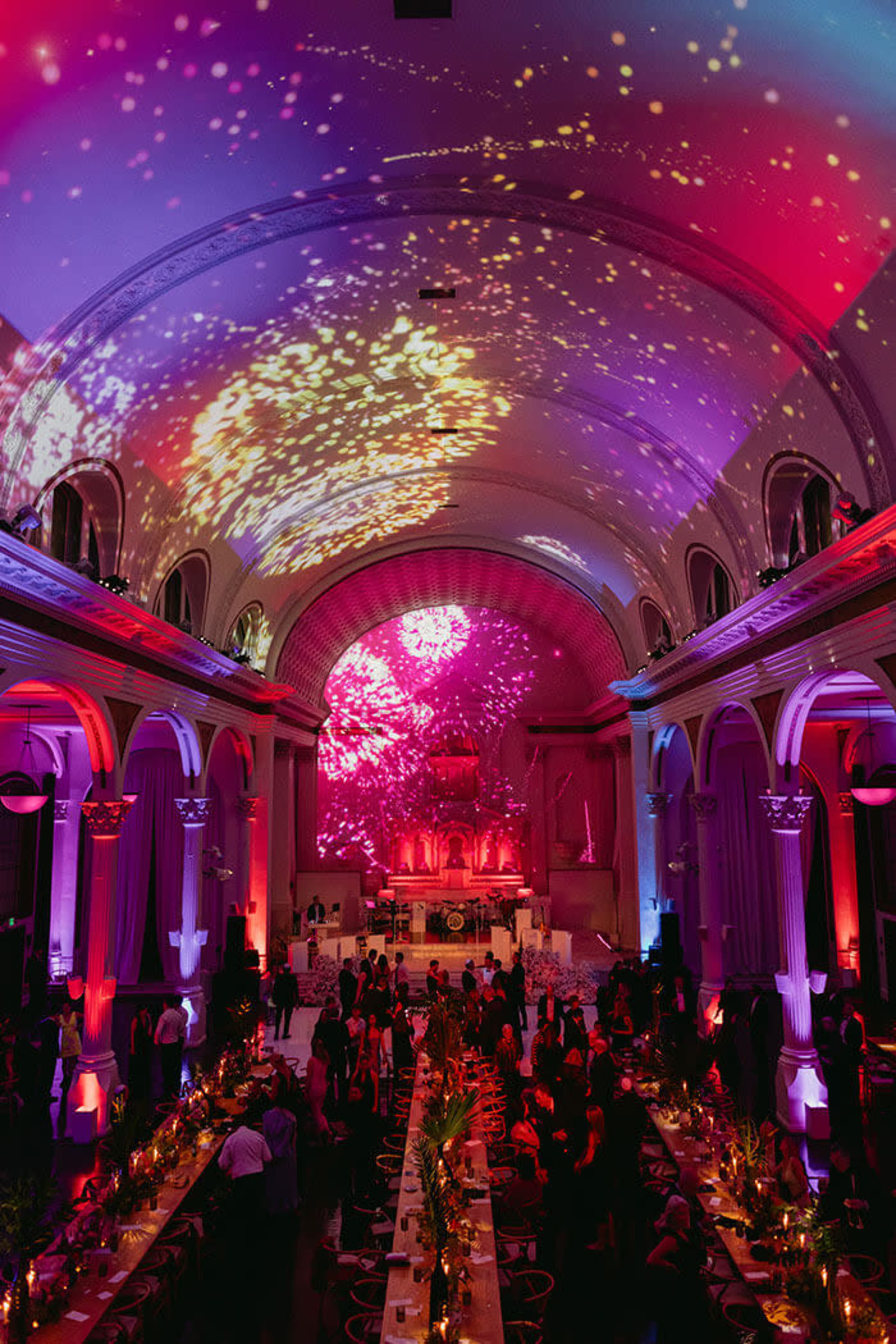 A large banquet hall is filled with guests, vibrant lighting casts colorful patterns on the ceiling, and a long dining table is set for an event.