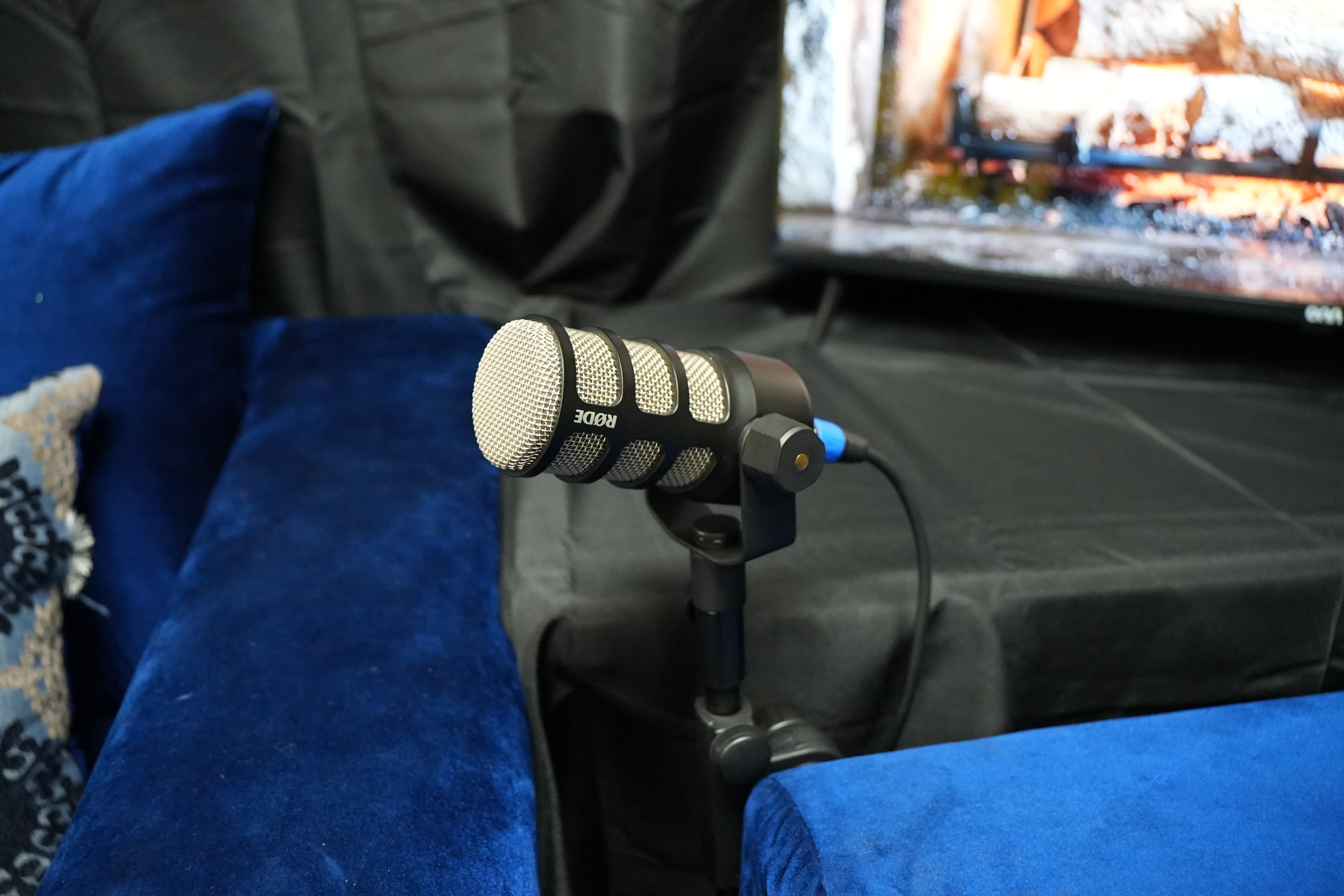 A microphone is positioned on a stand in front of a dark backdrop, with blue couches and a television displaying a fireplace scene.