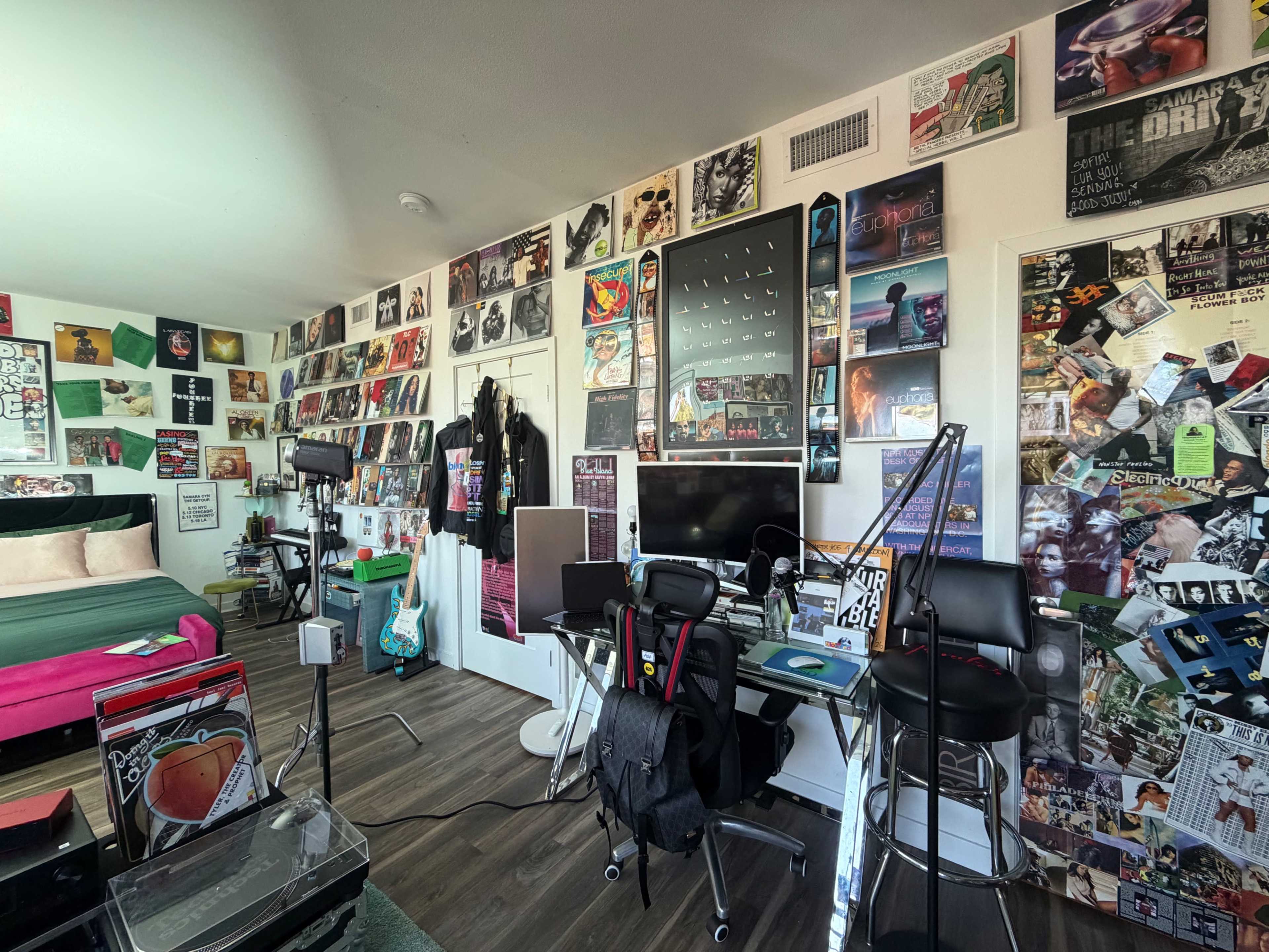 The image shows a room with a bed adorned with a pink comforter and a wall covered in numerous posters and photographs, alongside a desk area with two chairs and various music-related items.