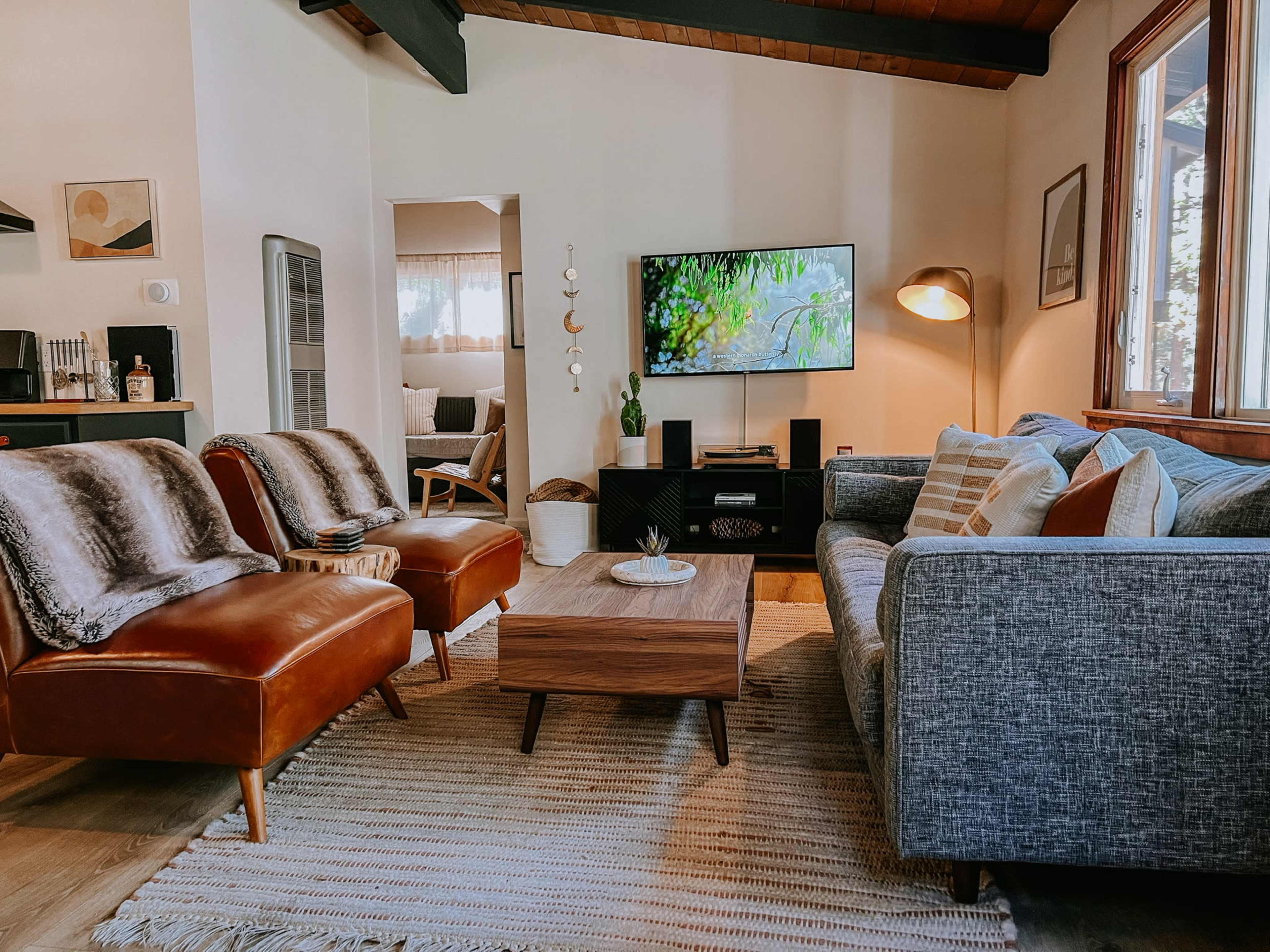 The image shows a cozy living room with two brown leather chairs, a gray sofa, a wooden coffee table, and a television displayed on the wall.