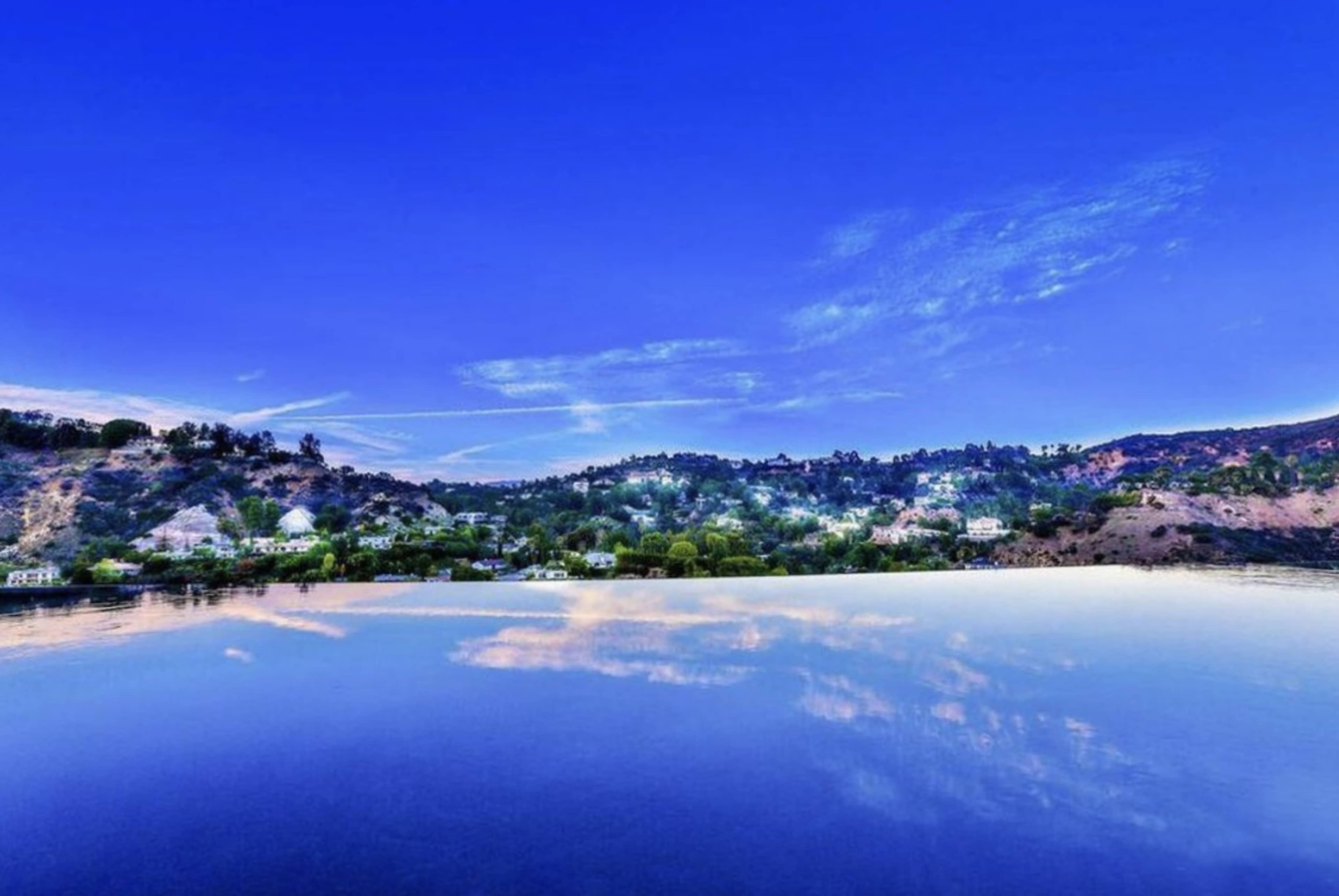 The image shows a calm body of water reflecting a clear blue sky and a hilly shoreline dotted with trees and homes.