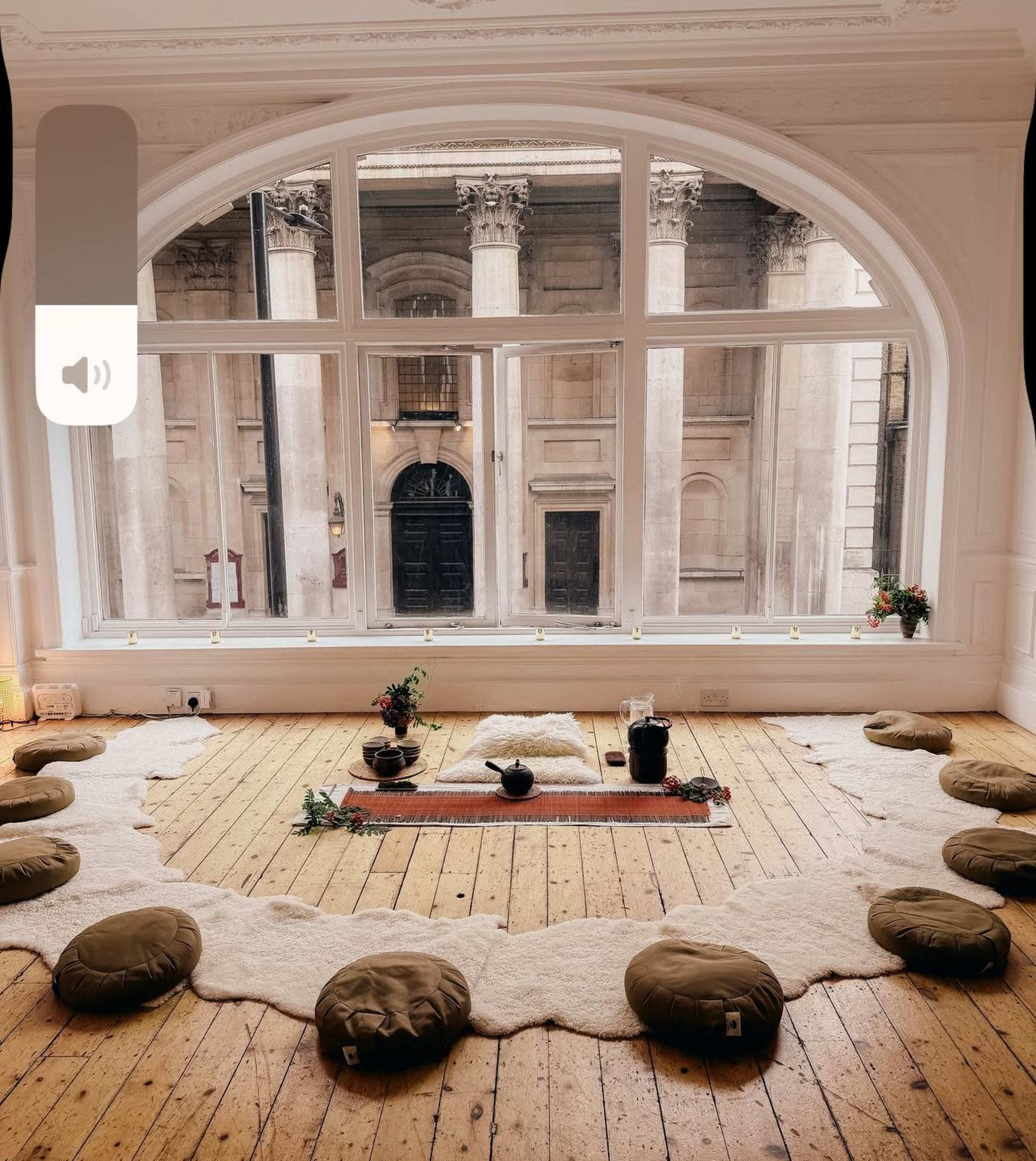 The image shows a spacious room with large arched windows, featuring a circular arrangement of floor cushions around a low table on a wooden floor.