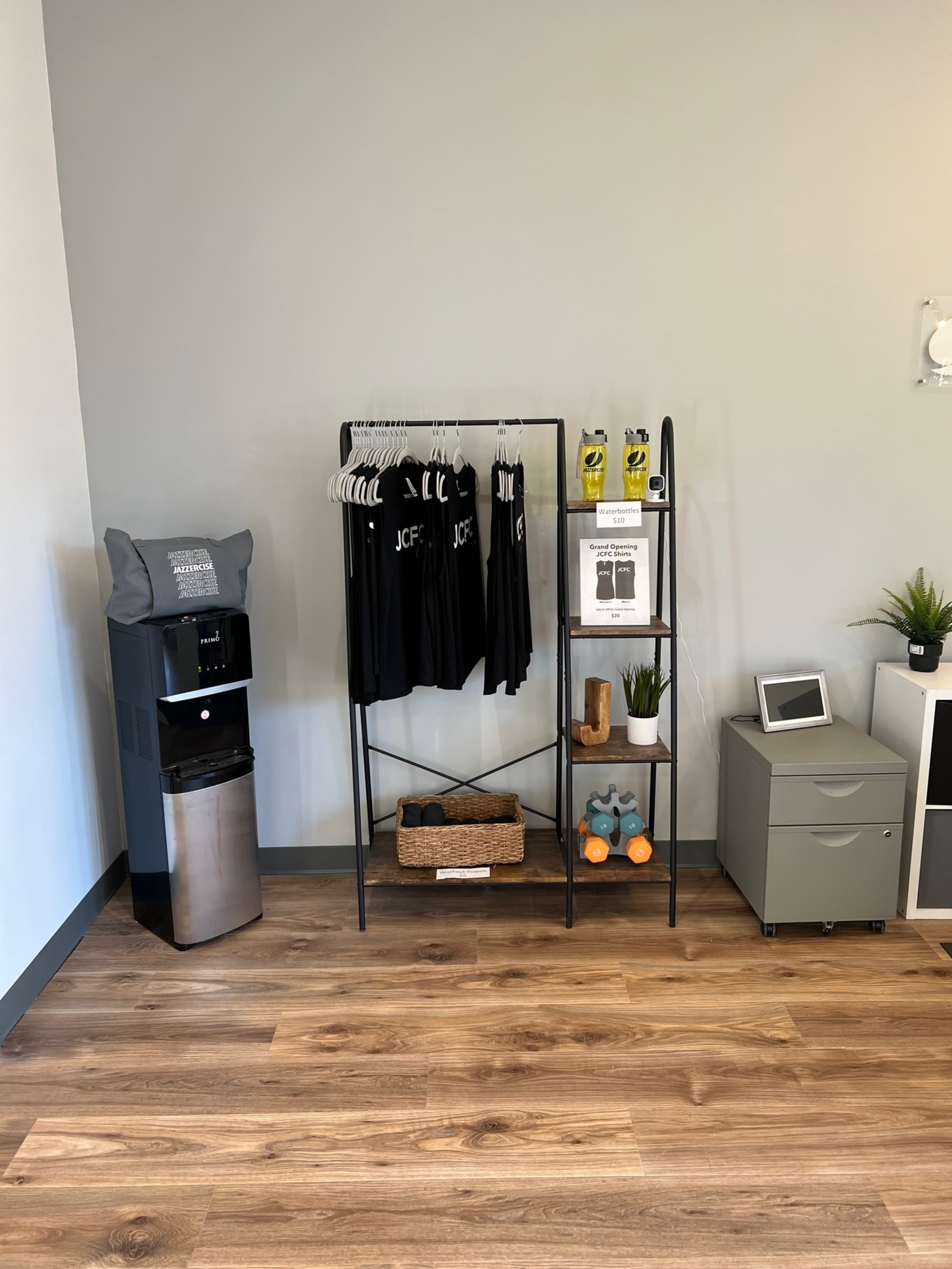 A neatly arranged interior with a clothing rack displaying black tops, a water cooler, a small shelf with drinks, and a smart tablet on a table against a gray wall.