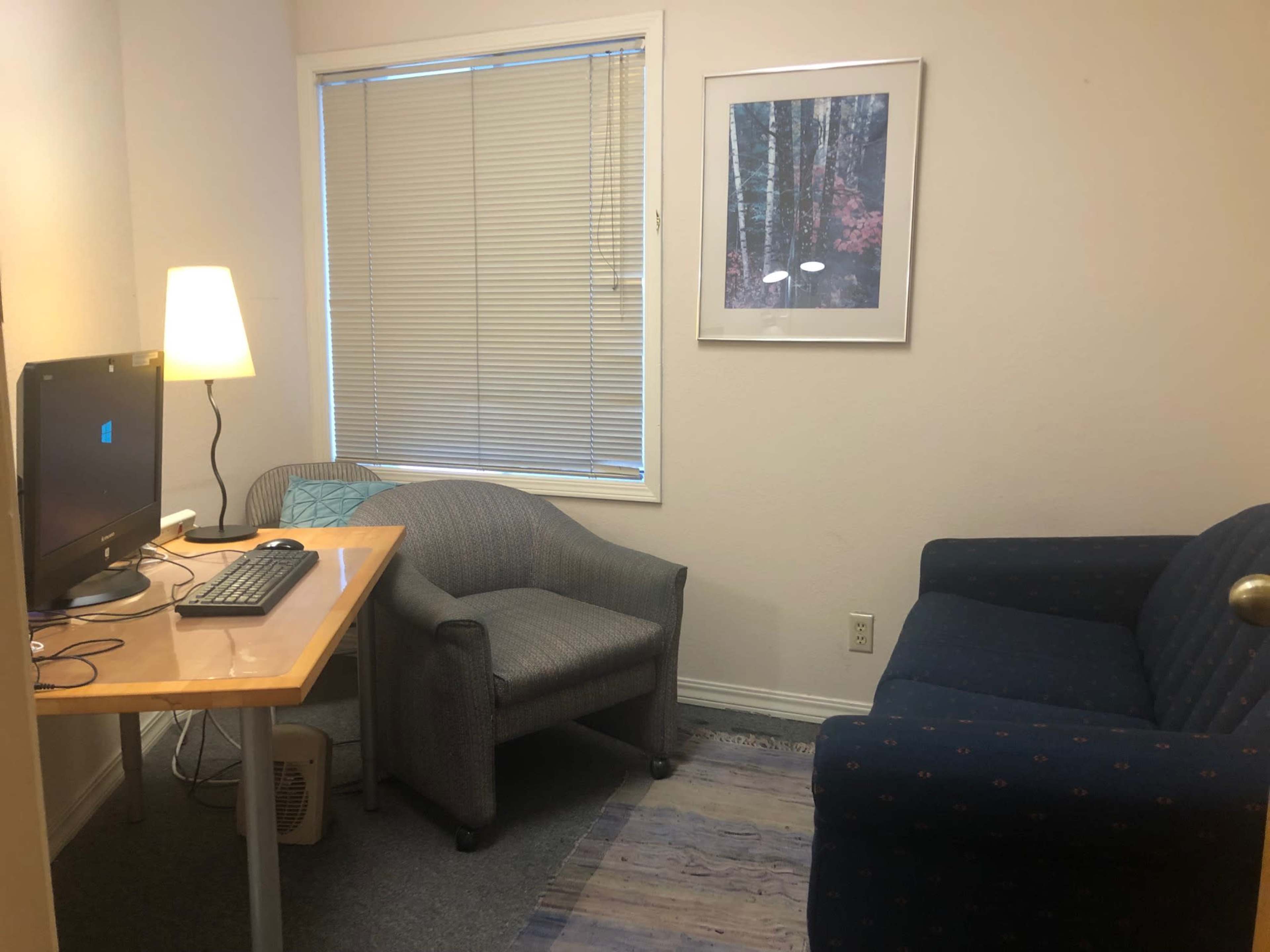The image shows a small room featuring a desk with a computer and a chair, a couch, and a window with blinds.