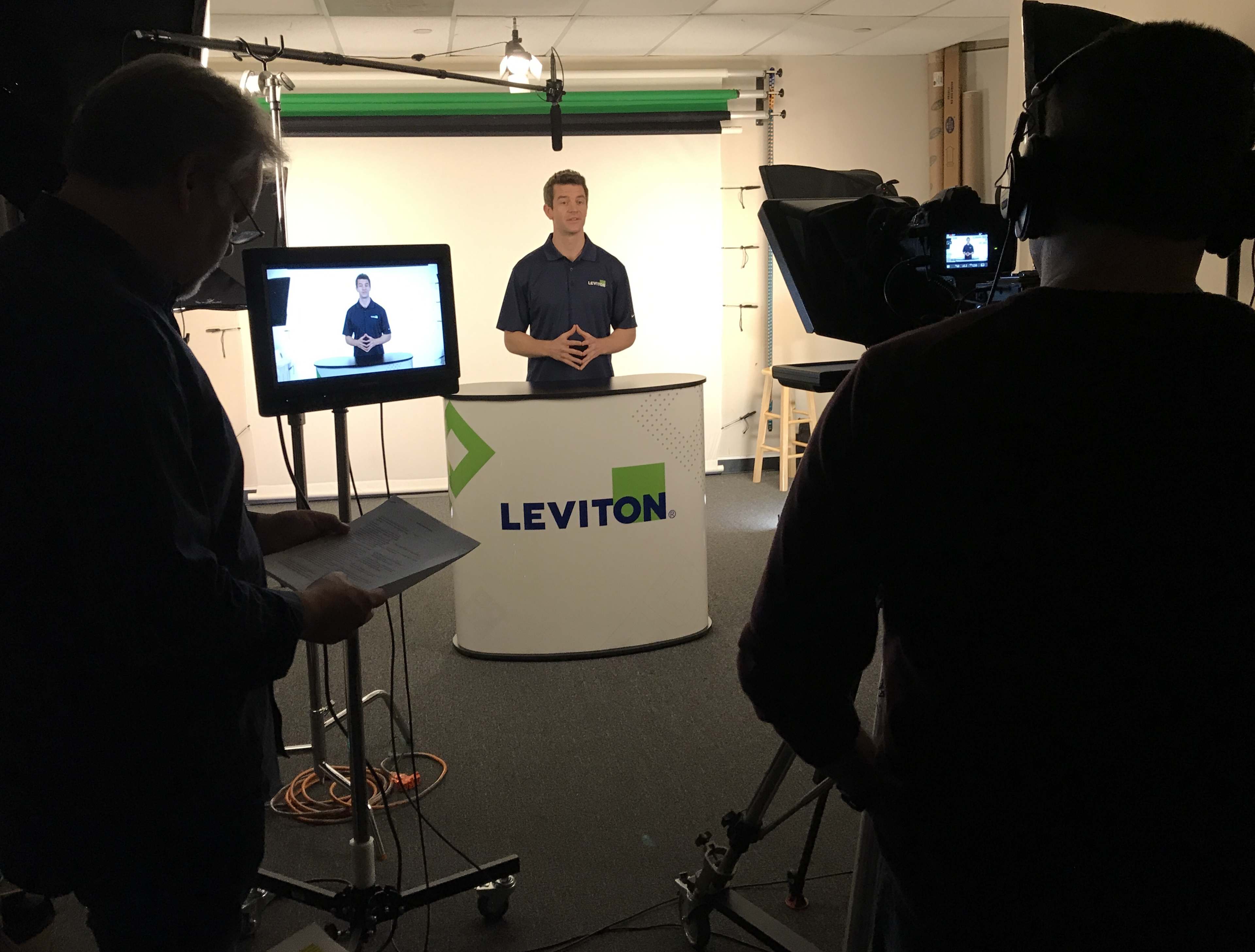 A person stands behind a display with the Leviton logo while another individual operates a camera and monitors the scene.