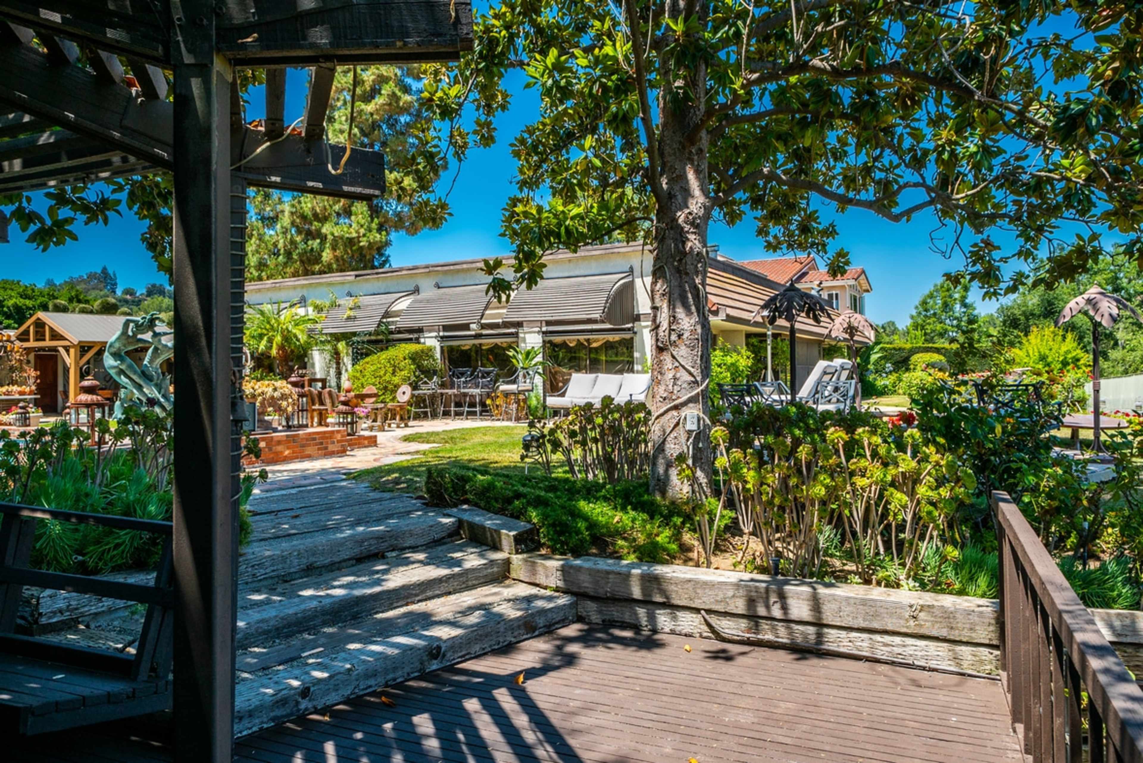 The image shows a well-maintained garden with a wooden deck, surrounded by greenery and a house with large windows in the background.