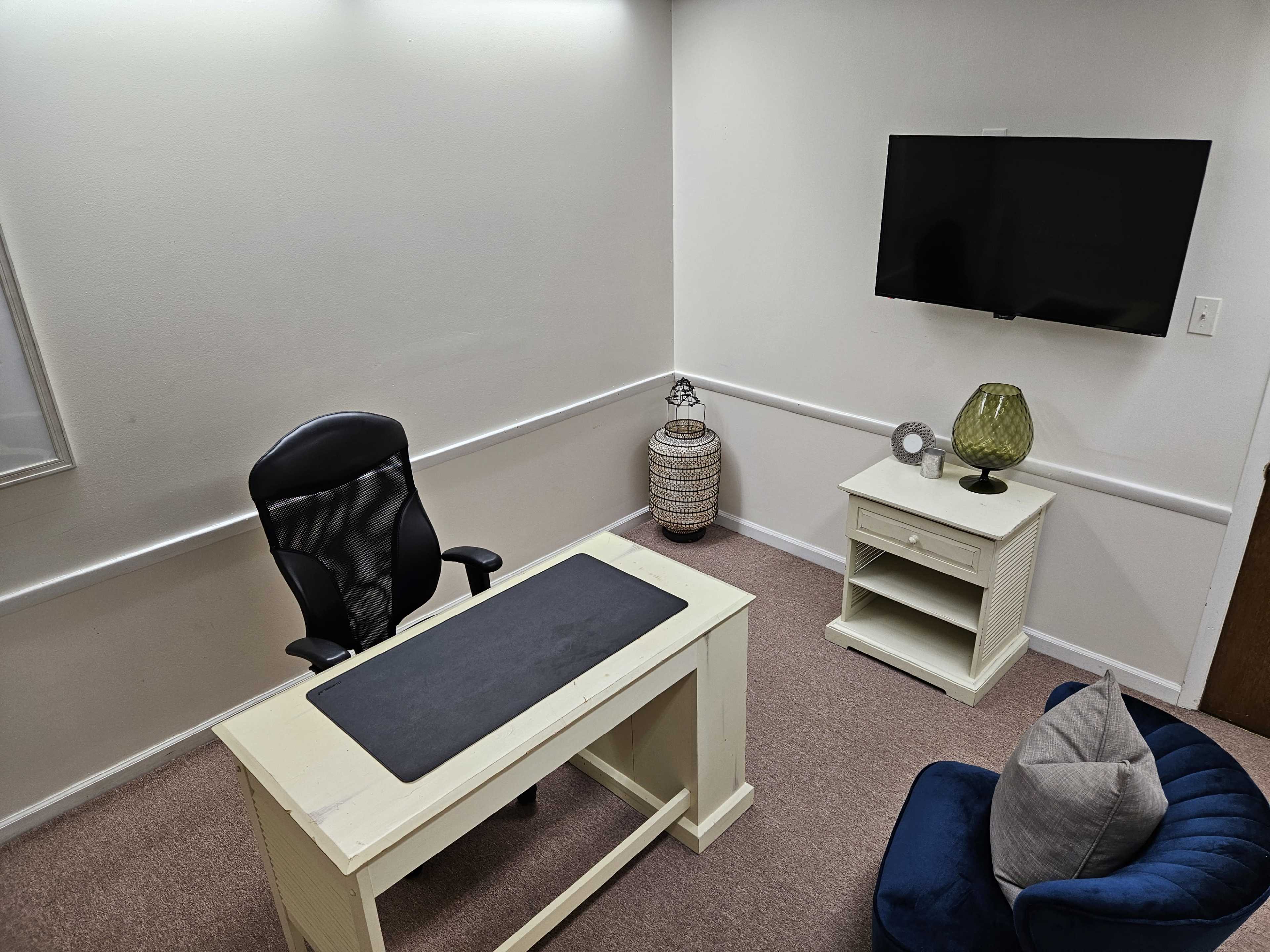 The image shows a small office space featuring a desk with a black chair, a television mounted on the wall, a side table, and a decorative lamp in the corner.