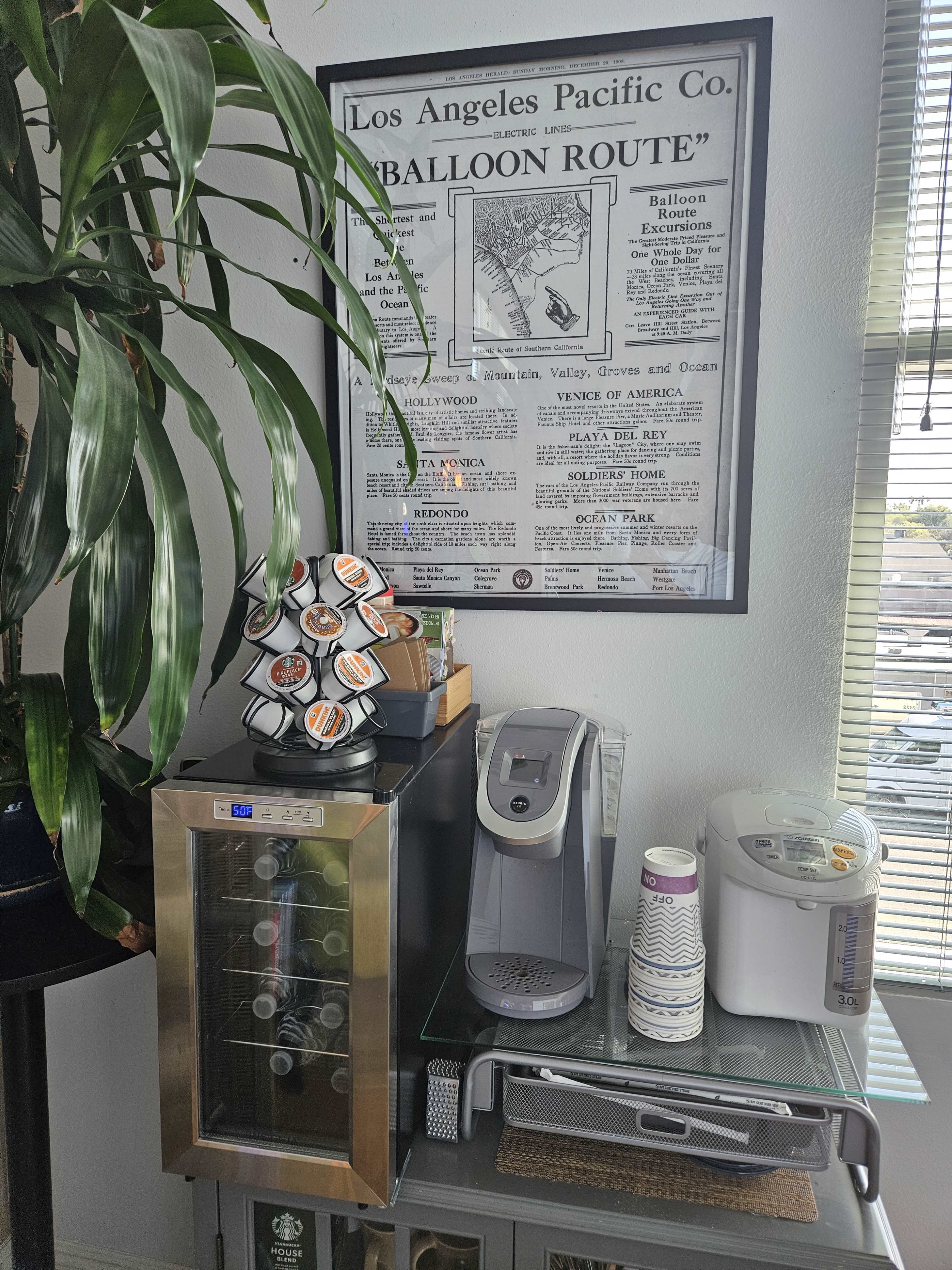A small beverage station featuring a wine cooler, a coffee maker, and other drink-related items next to a large framed vintage advertisement.