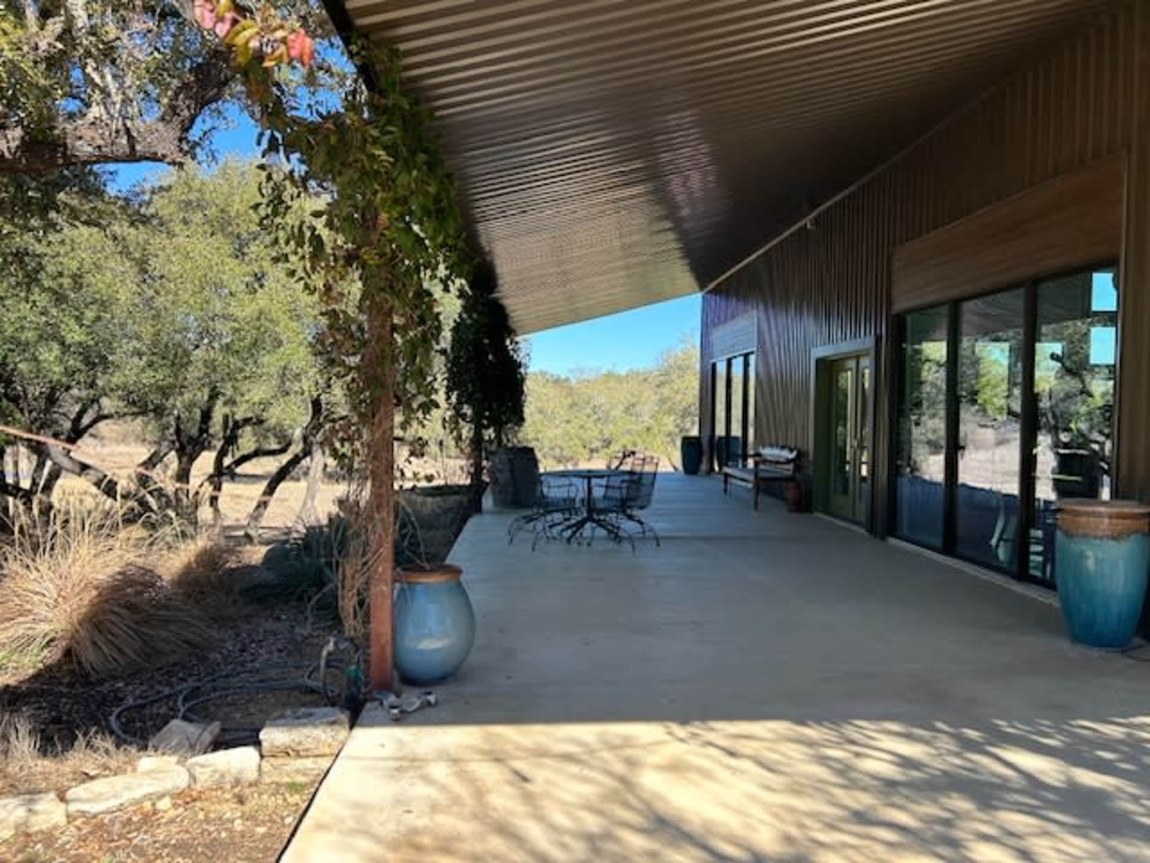 The image shows a spacious concrete patio with metal roofing, surrounded by trees and open land, featuring a table and chairs, along with decorative planters.