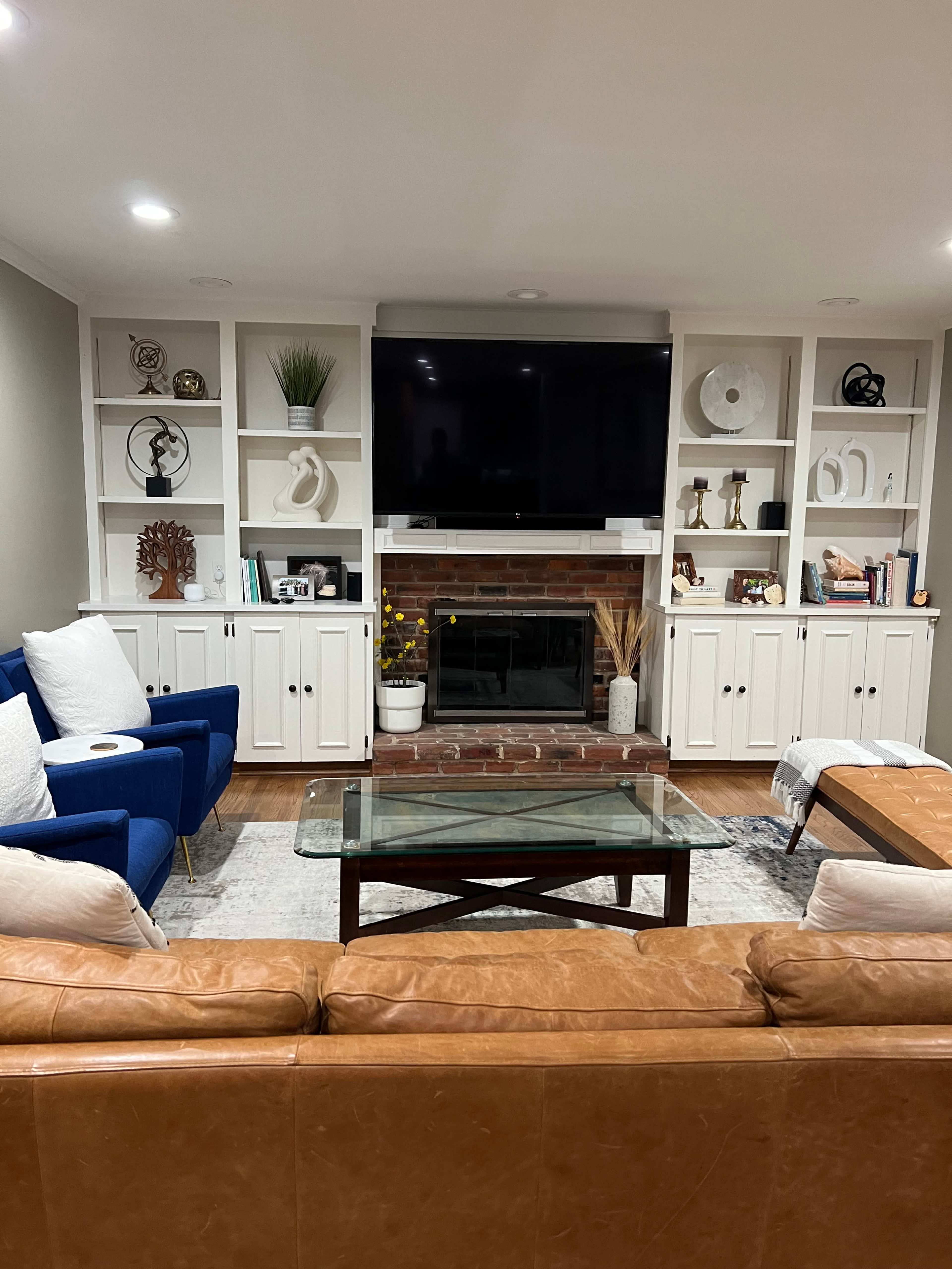 A living room features a brick fireplace surrounded by built-in shelves, with a large television mounted above, two blue chairs, a brown leather sofa, and a glass coffee table.
