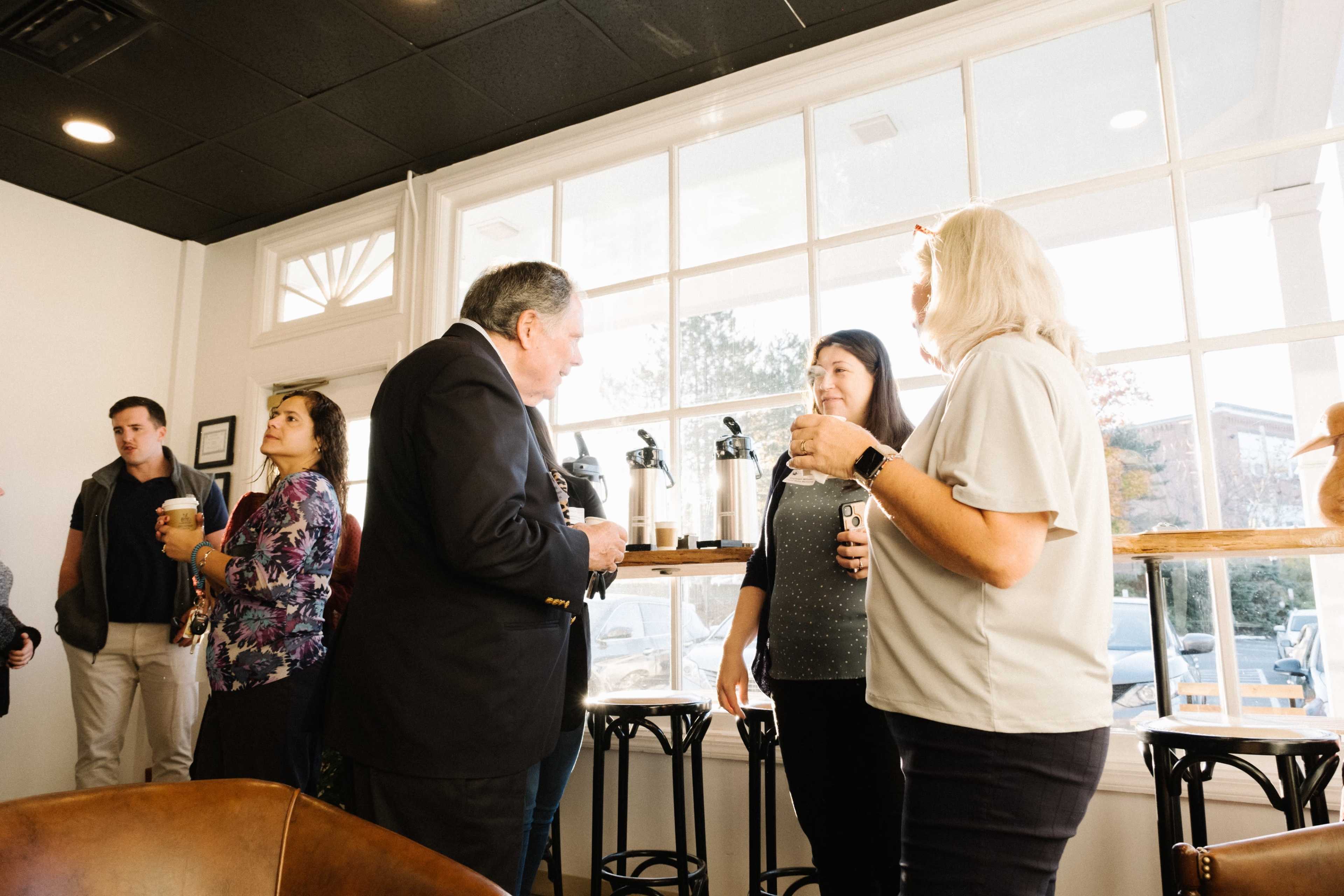 A group of people engages in conversation near a window in a café, with coffee dispensers visible in the background.