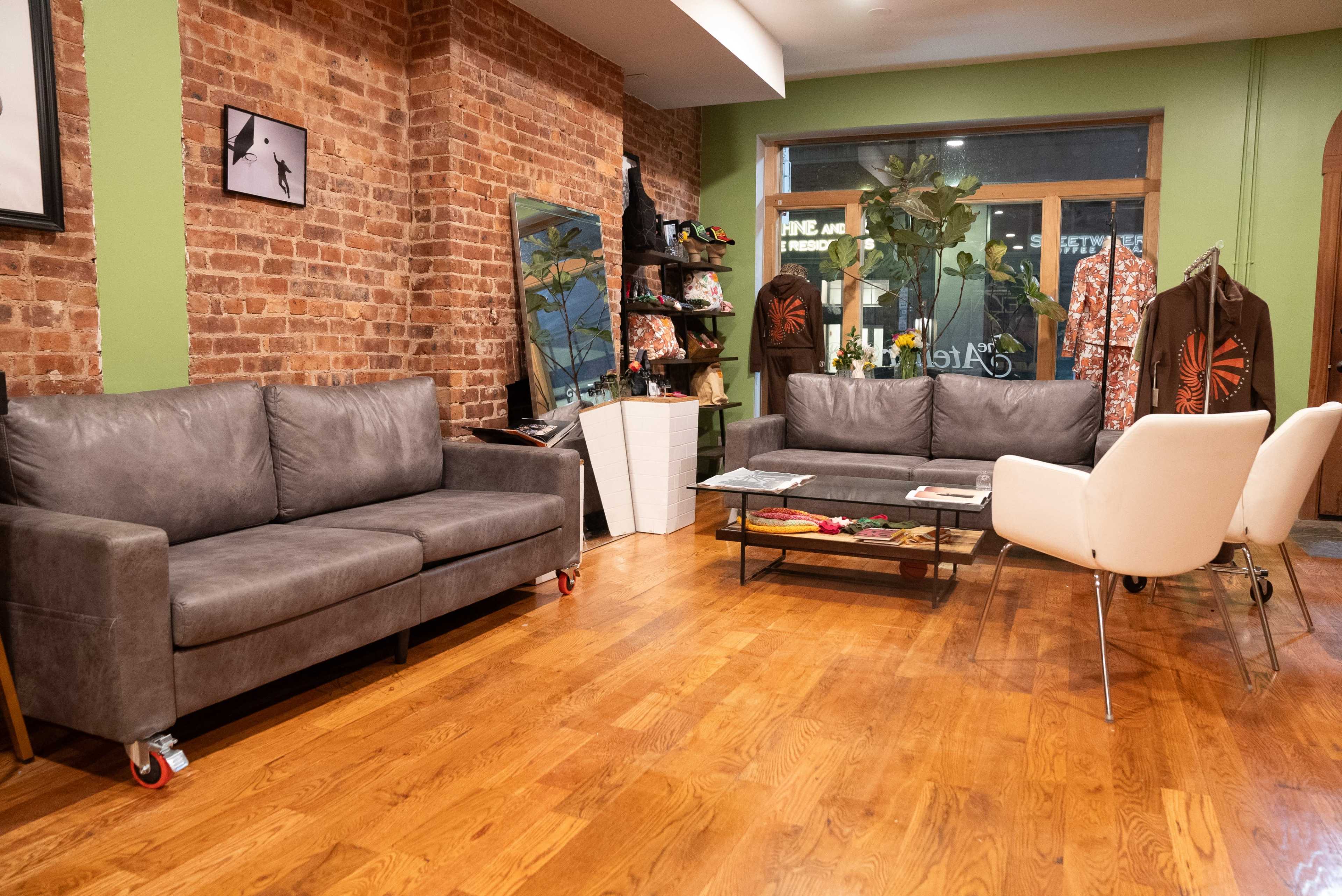 The image shows a cozy interior with two gray sofas, a white chair, a wooden floor, and brick walls, featuring shelves with decorative items and mirrors.