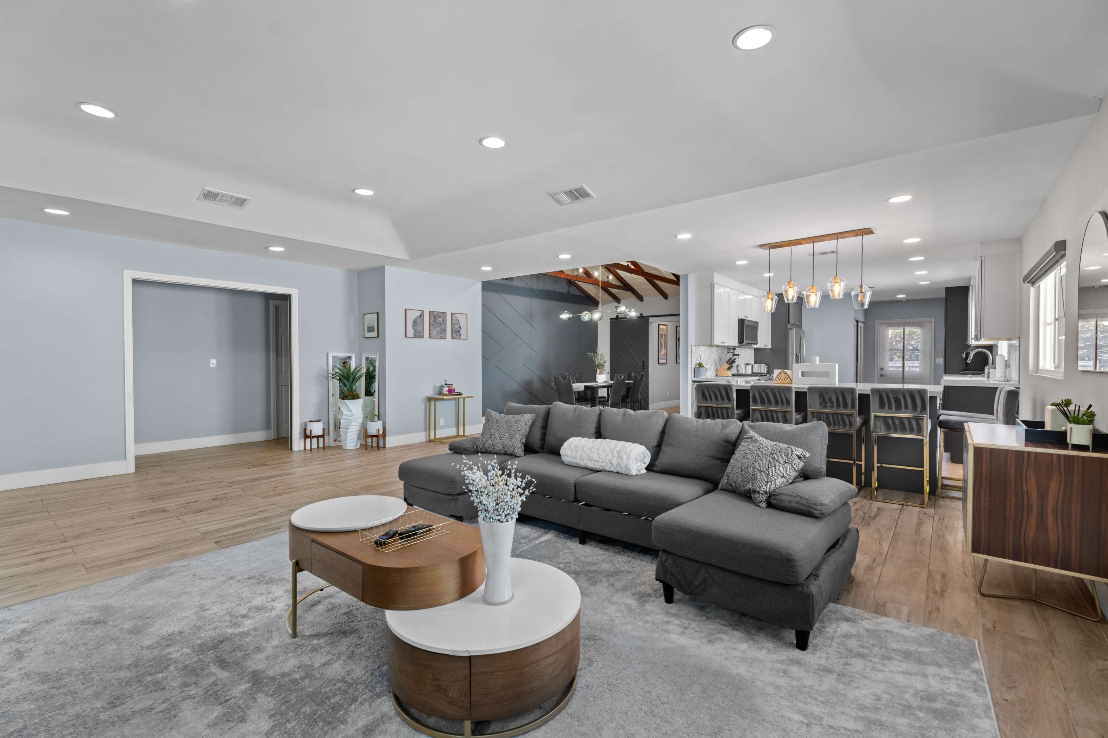 A spacious living room features a gray sectional sofa, a round coffee table, and a view into a modern kitchen equipped with an island and dining area.