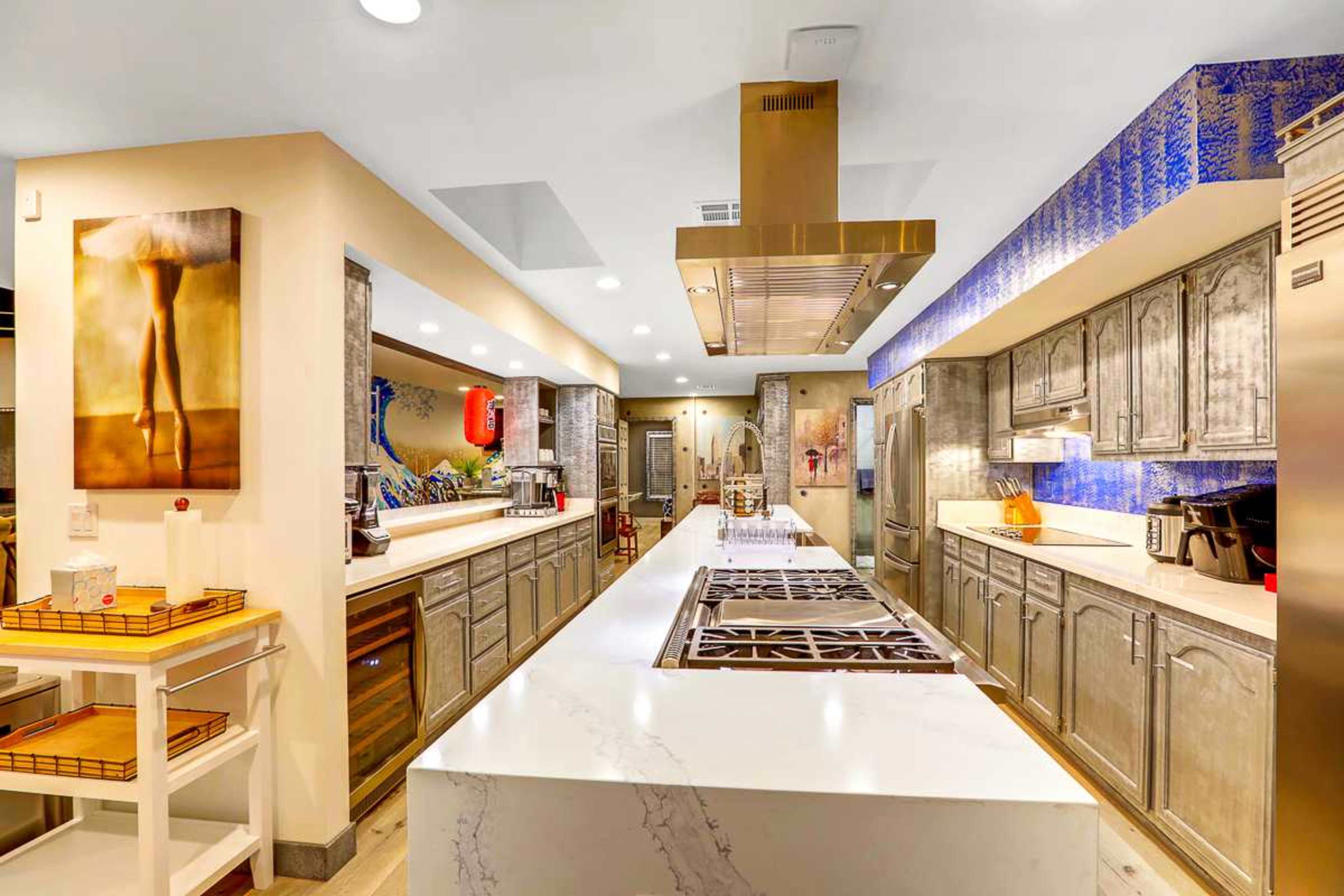 The kitchen features a large central island with a stovetop, stainless steel appliances, and metallic cabinets against a blue backed wall.