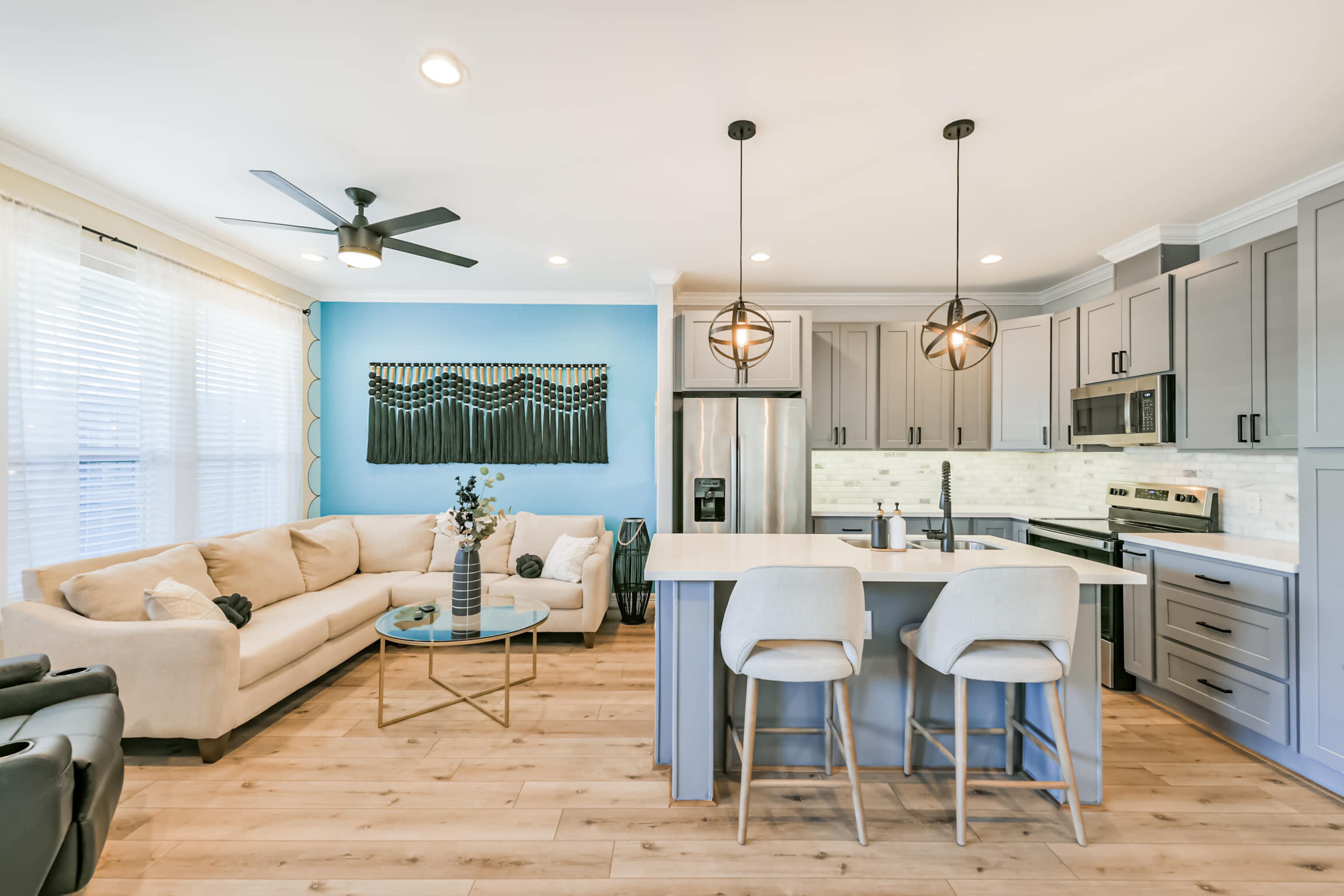 The image shows a modern living room and kitchen area with a beige sofa, circular glass table, and gray cabinetry.