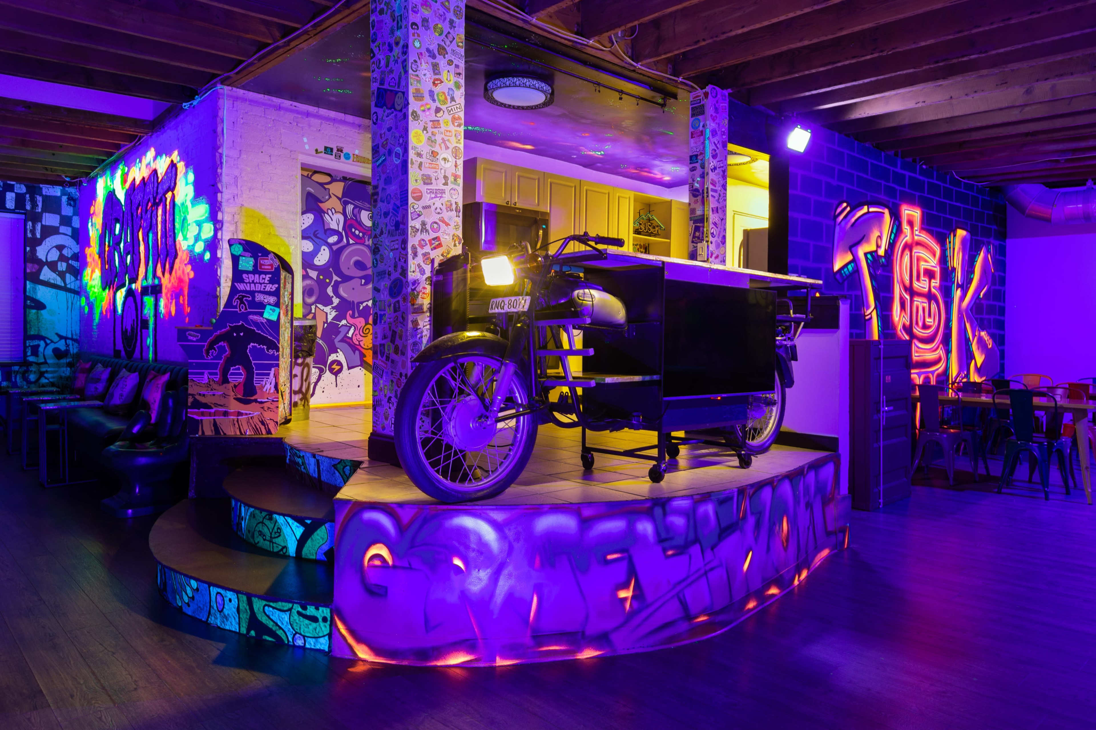 The image shows a colorful, artfully decorated space featuring a prominent motorcycle delivery cart on a raised platform, illuminated with vibrant UV lighting.