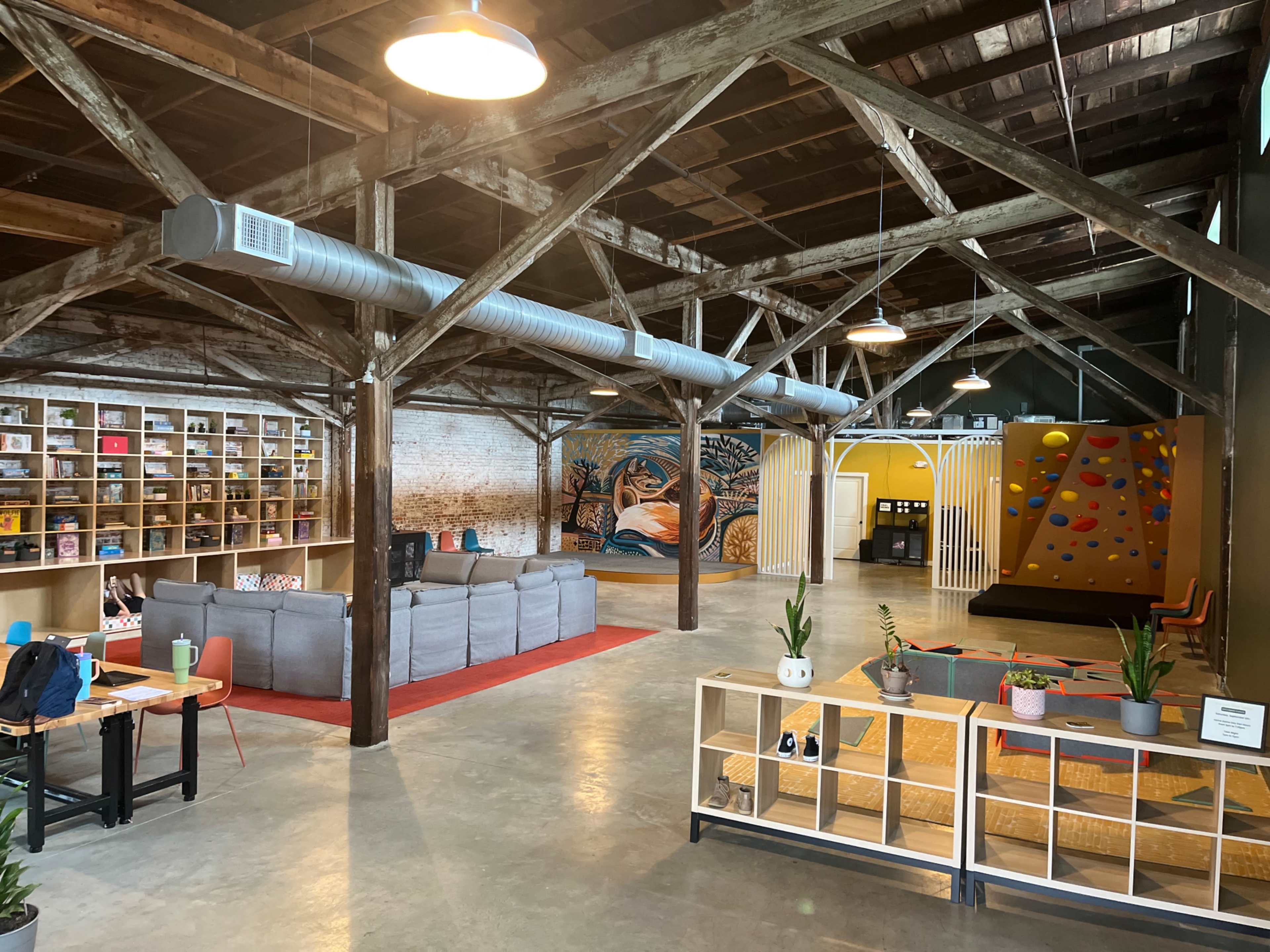 The image shows an industrial-style interior with exposed beams, featuring a seating area, bookshelves, a colorful mural, and a climbing wall.