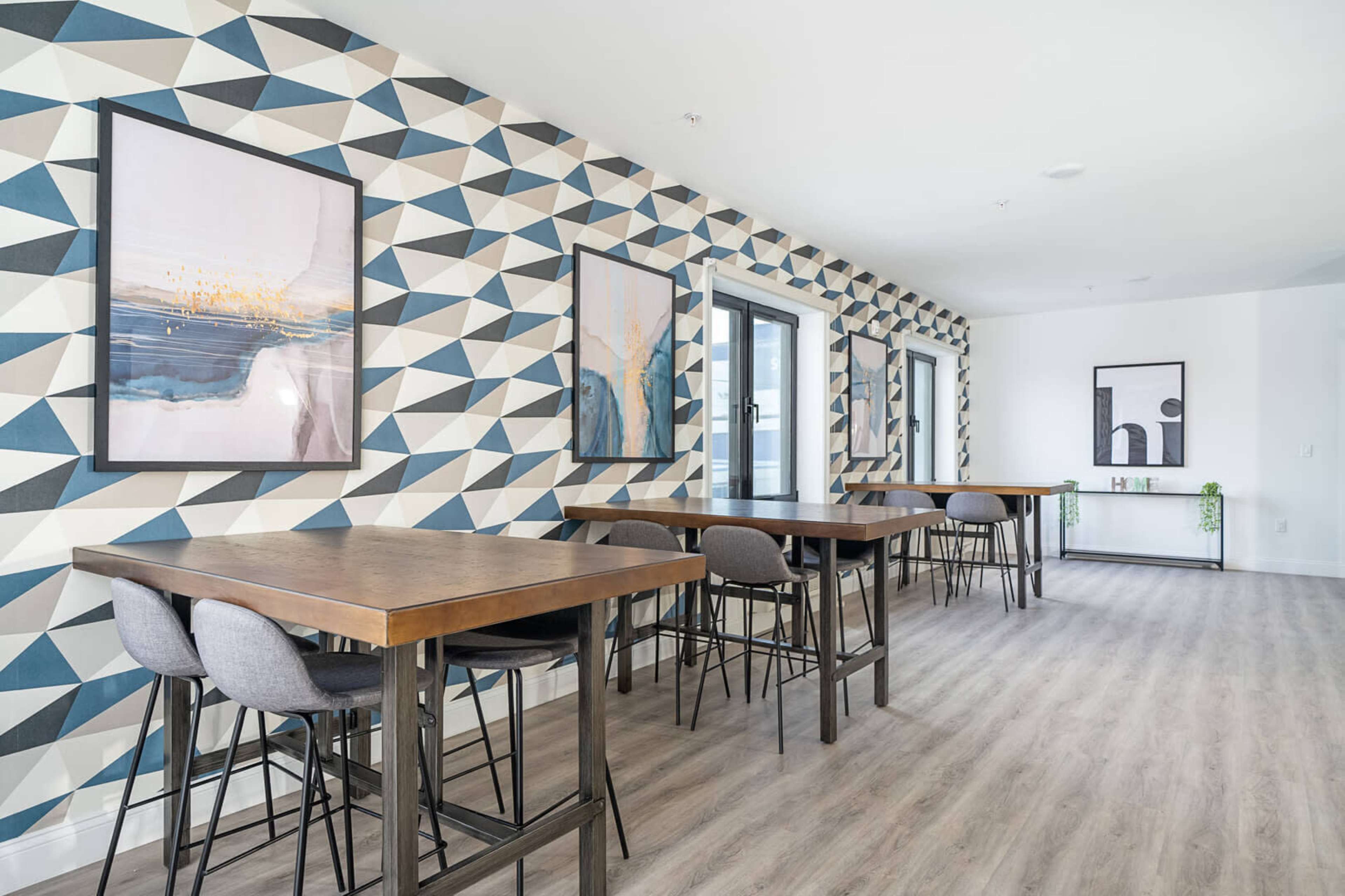 A modern room featuring two rows of tall wooden tables with bar stools, set against a wall adorned with a geometric patterned wallpaper in blue and neutral tones.