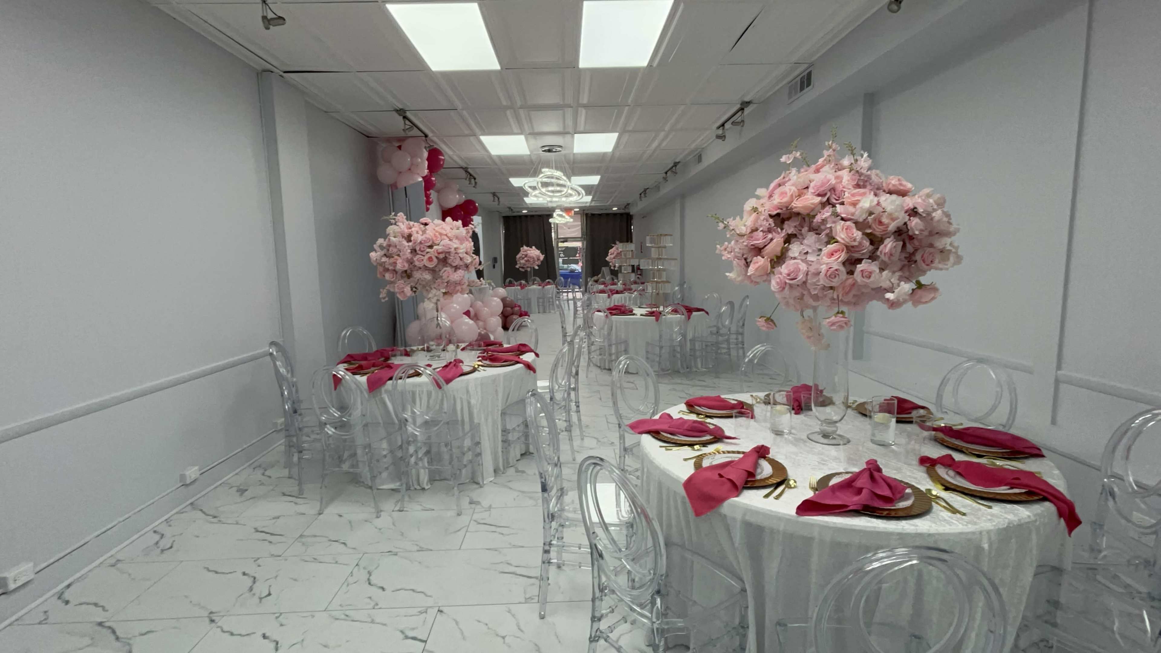 The image shows an elegantly decorated event space with round tables covered in white cloth, clear chairs, and centerpieces featuring large pink floral arrangements.