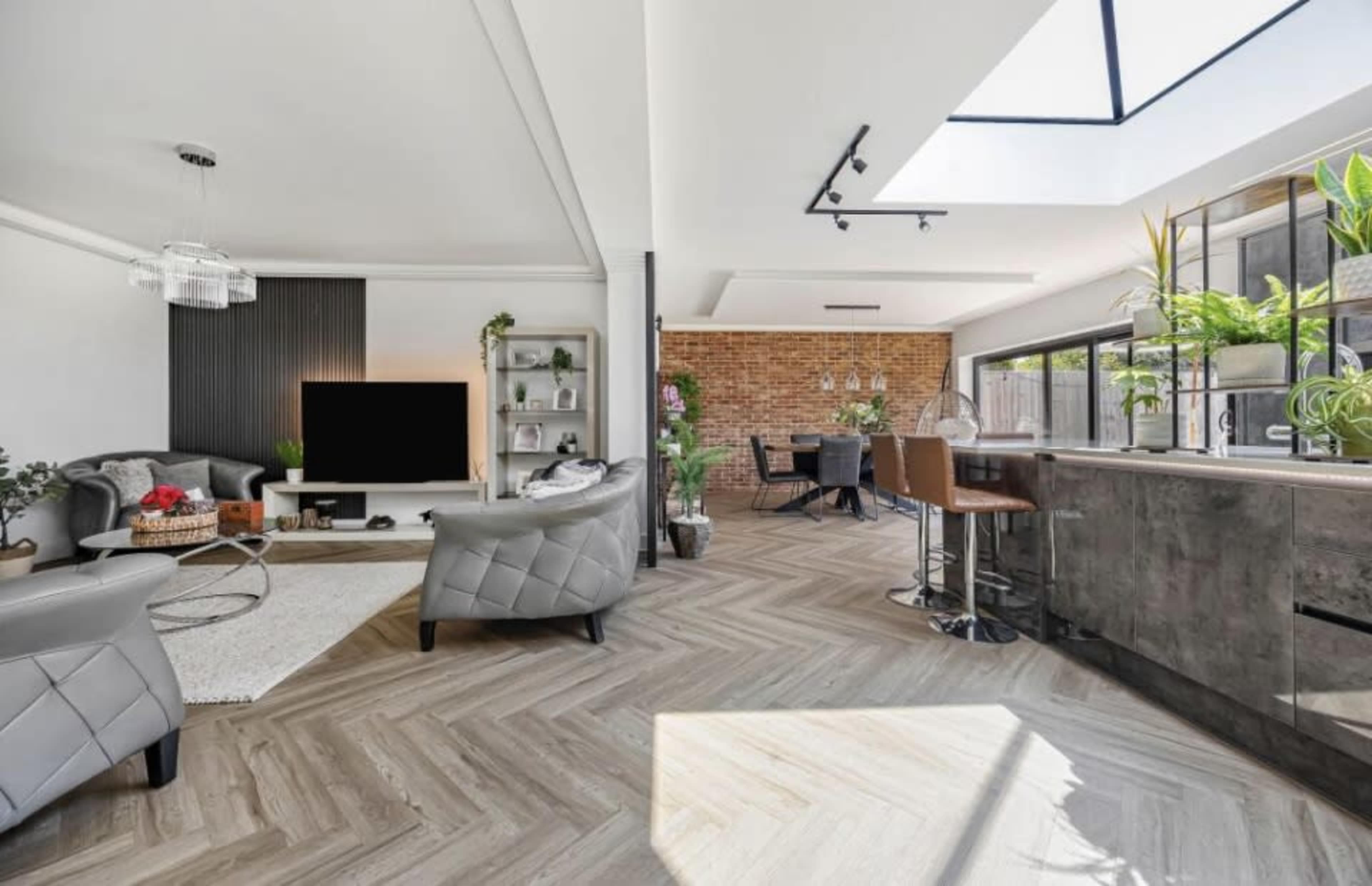 The image shows a modern open-concept living space with a grey couch, a television, a dining area, and large windows that allow natural light to flood the room.