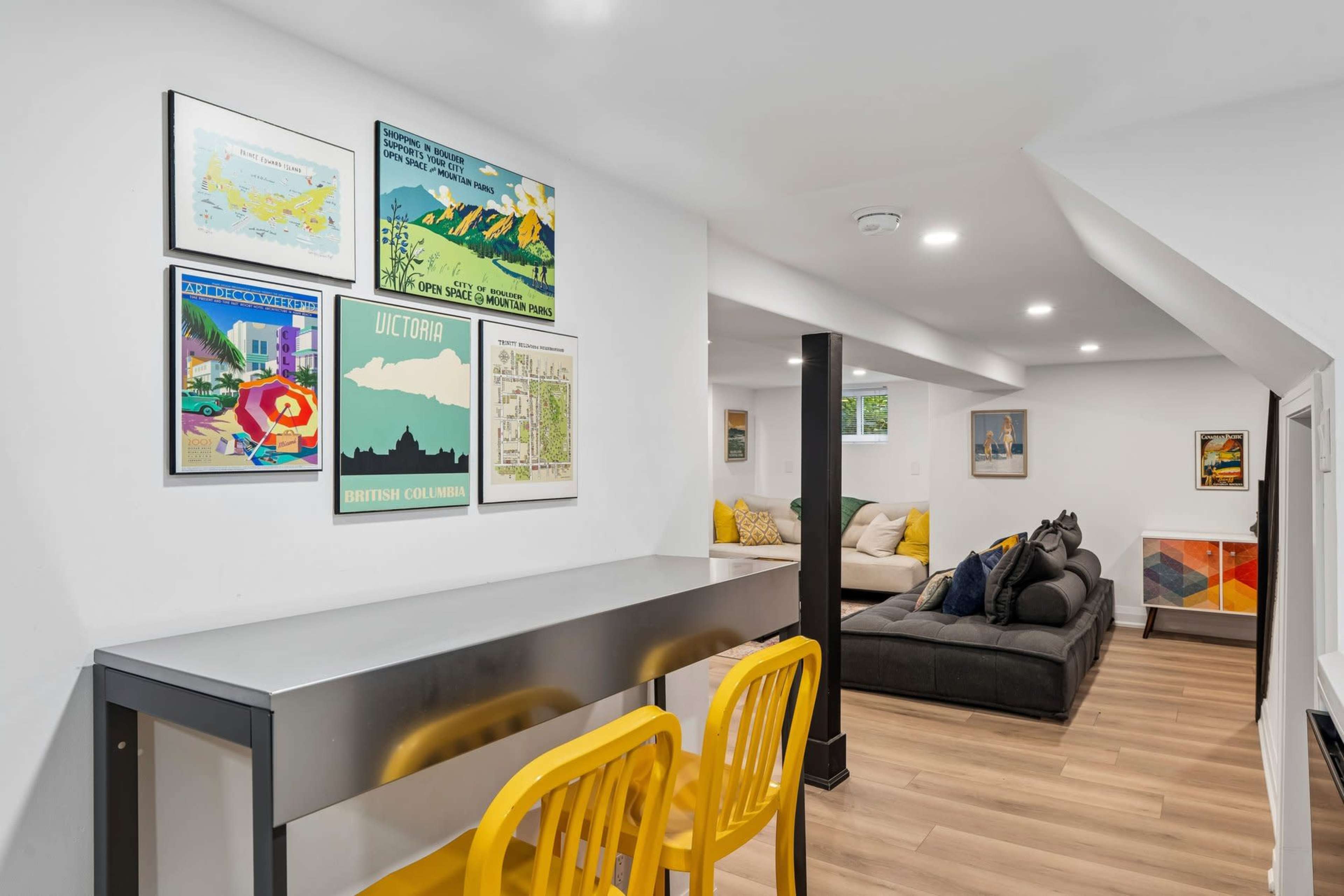 A light-colored interior features a small dining area with yellow chairs and a metallic table, while a cozy living space with a sectional sofa and artwork is visible in the background.