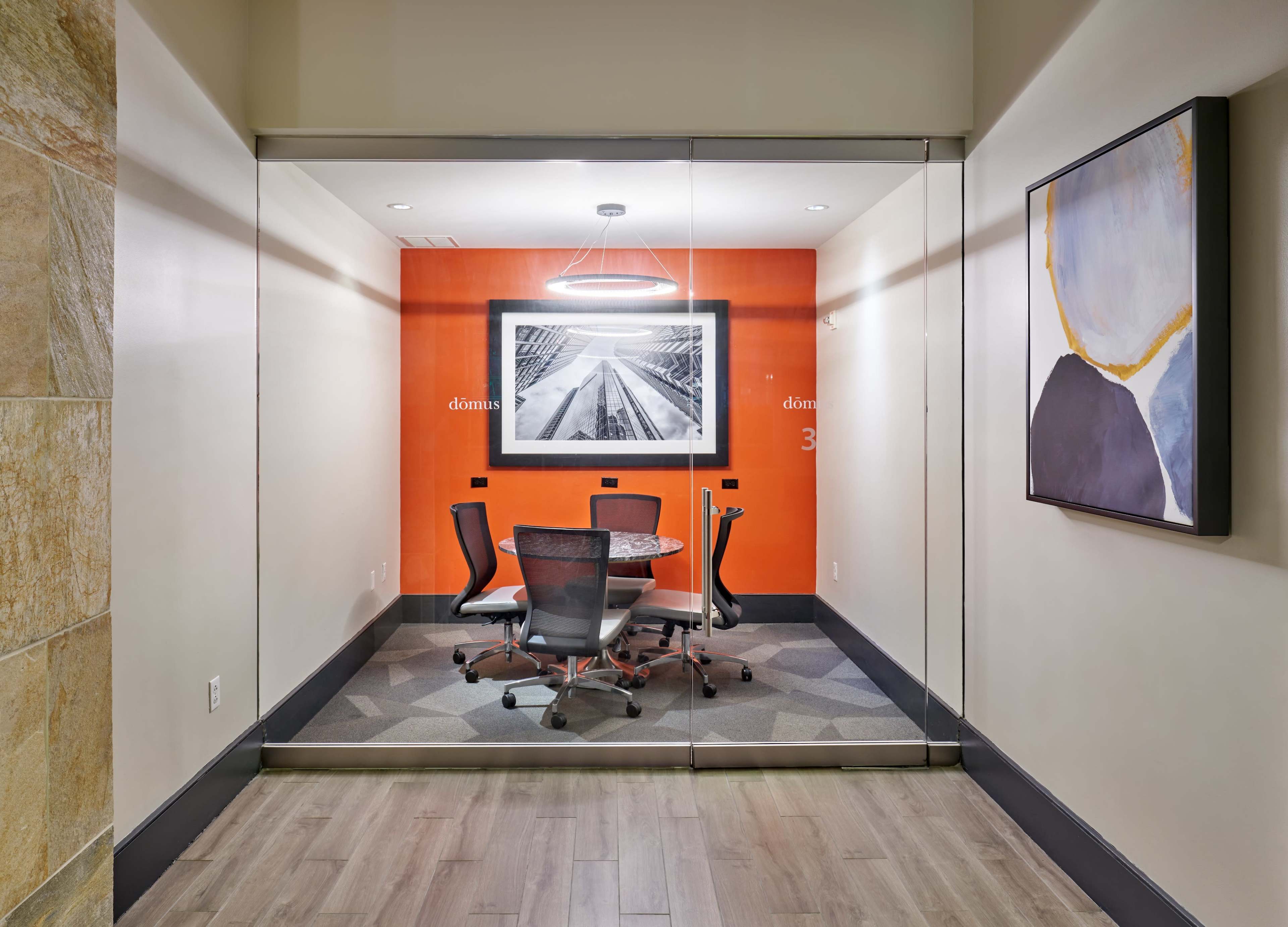 The image shows a modern conference room surrounded by glass walls, featuring a round table and four chairs against an orange accent wall adorned with a black and white photograph.