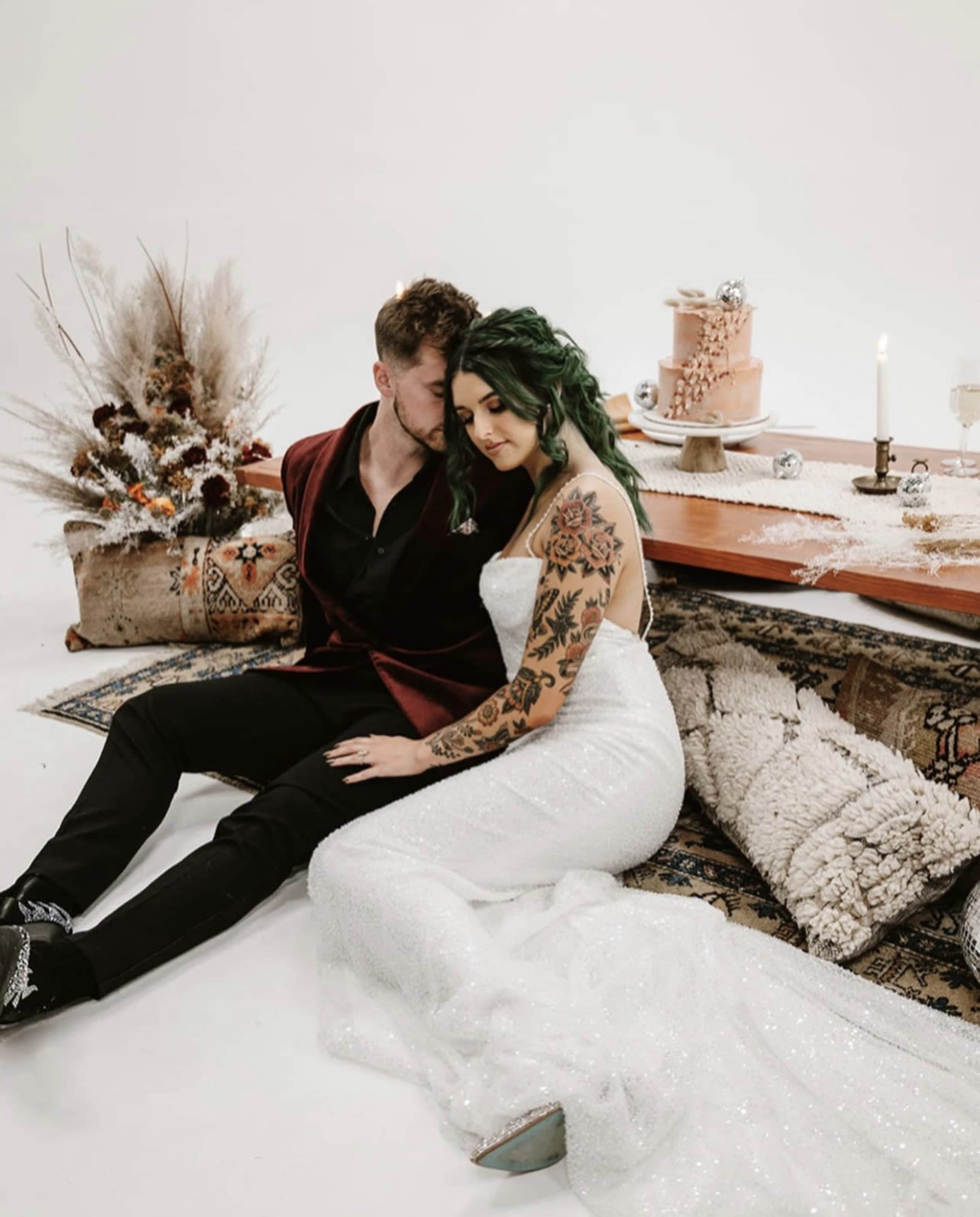 A couple sits on the floor next to a table set with a cake and decorative elements, surrounded by a rustic arrangement of plants and textiles.