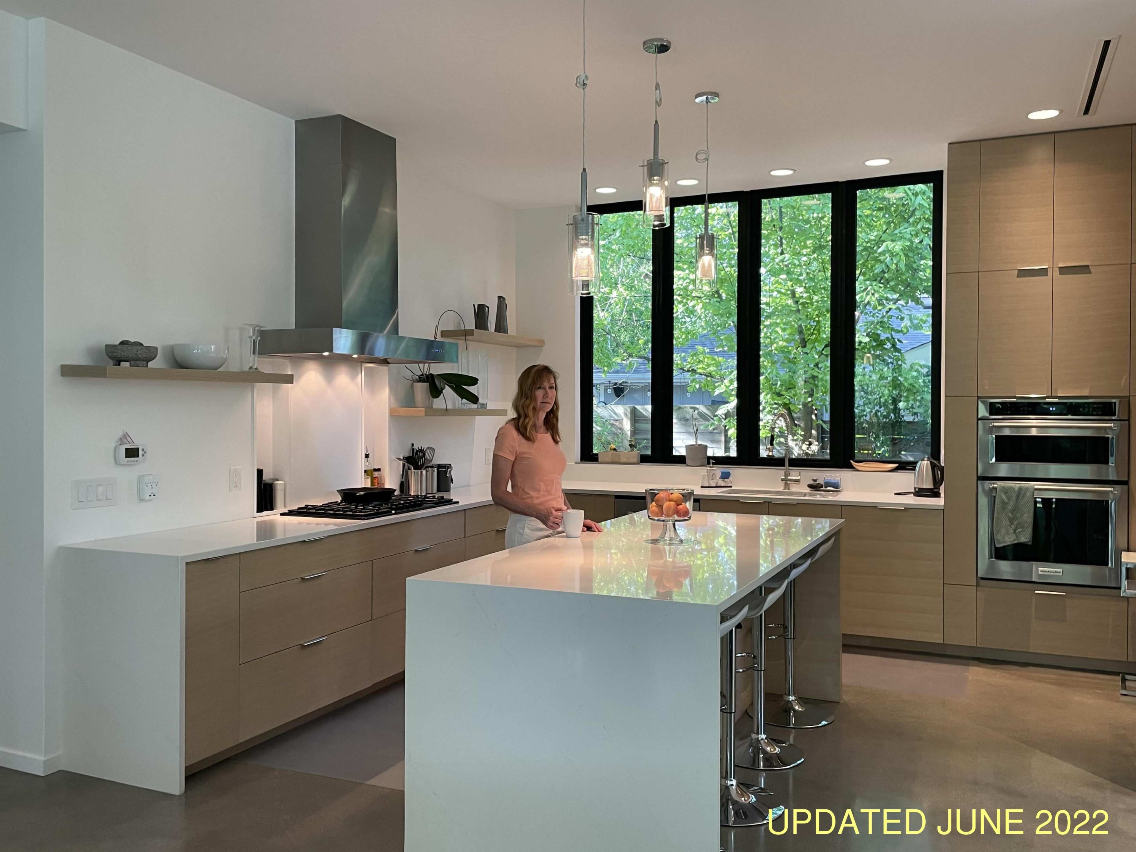A woman stands in a modern kitchen with large windows overlooking greenery, featuring sleek cabinets and an island with bar stools.