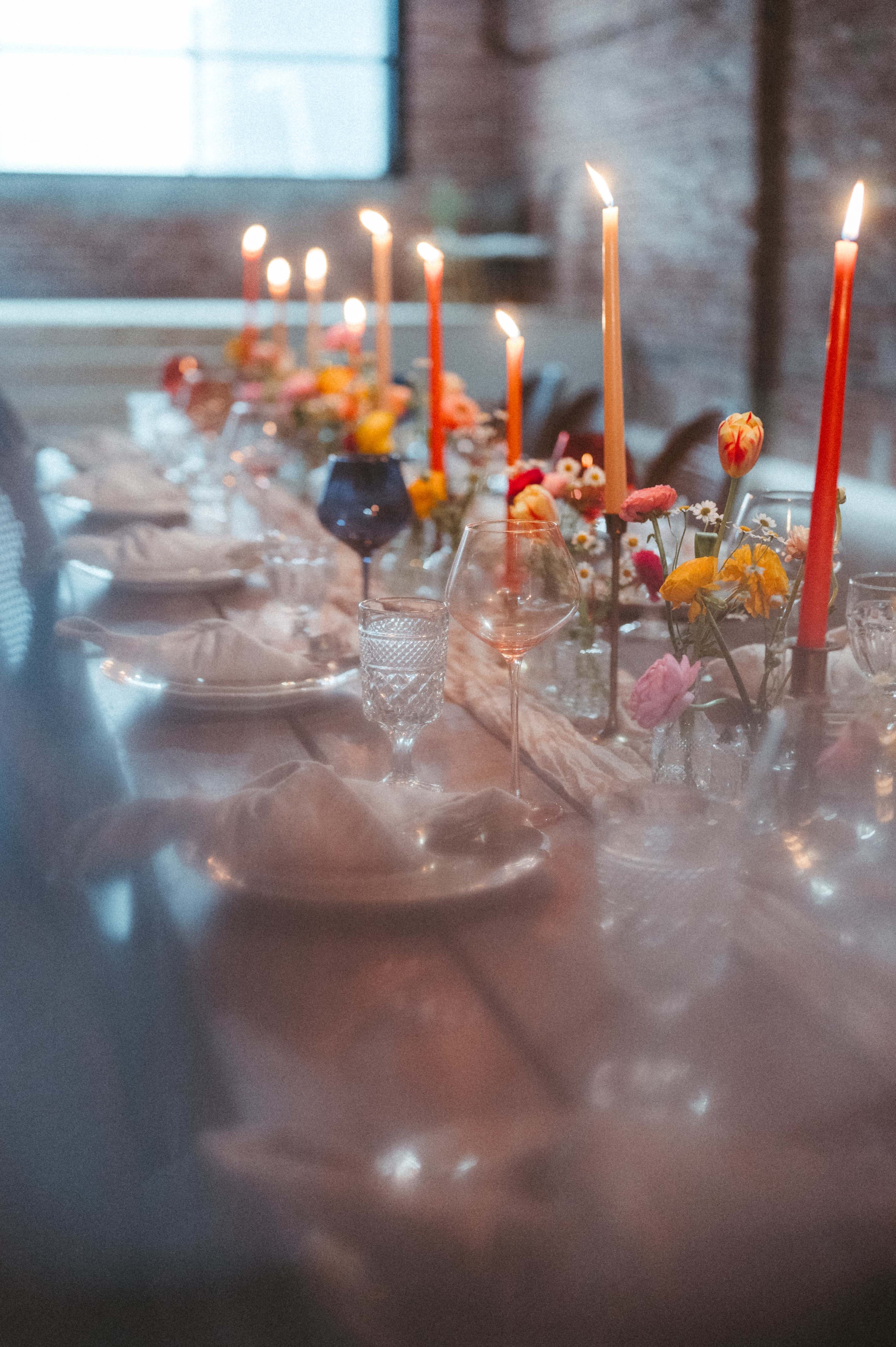 A long dining table is set with plates, crystal glasses, and colorful flowers, surrounded by lit candles in various heights.