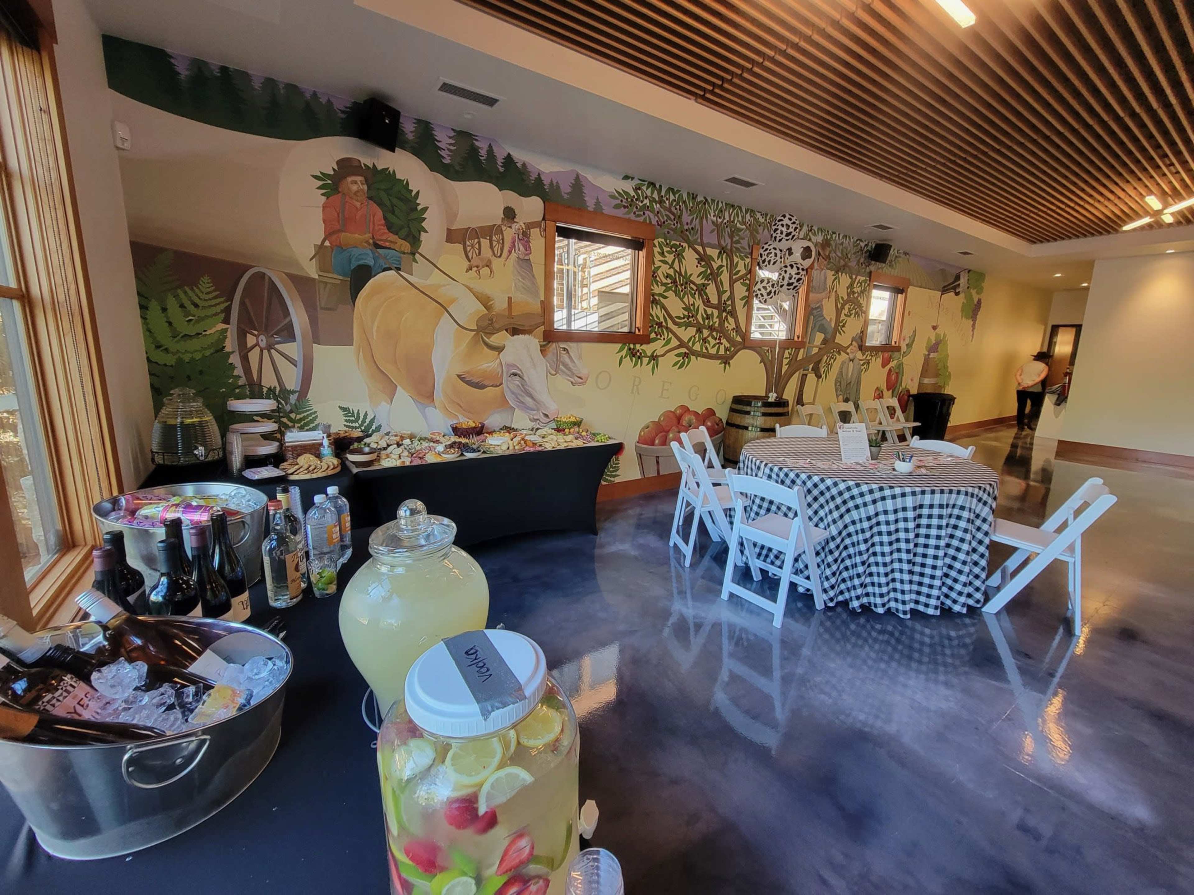 A spacious indoor event space with a mural depicting a farmer, a table laden with food, and a drink station featuring a large glass jar of lemonade.