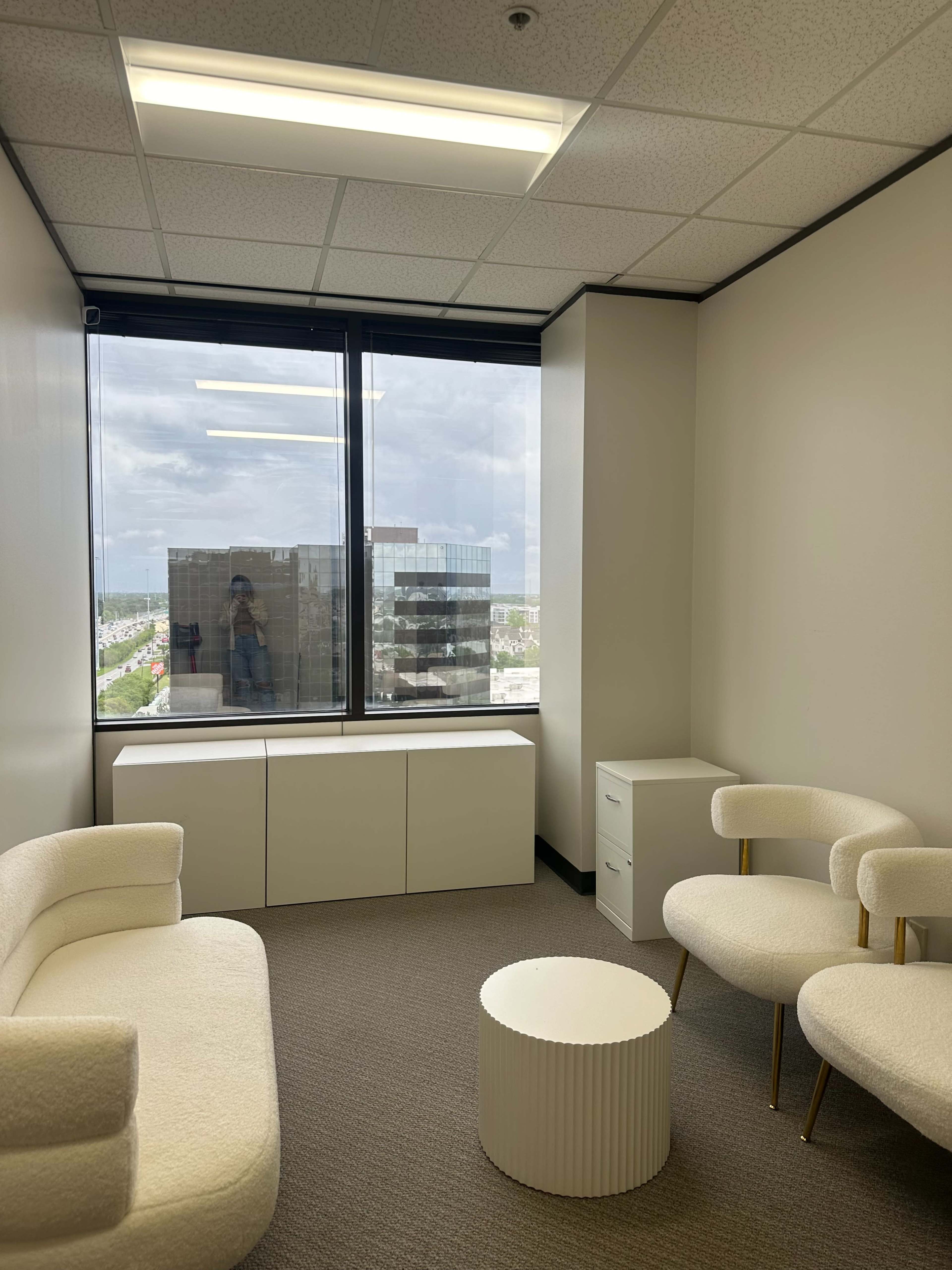 A small office space features two chairs, a circular table, and a large window with a view of the city skyline.