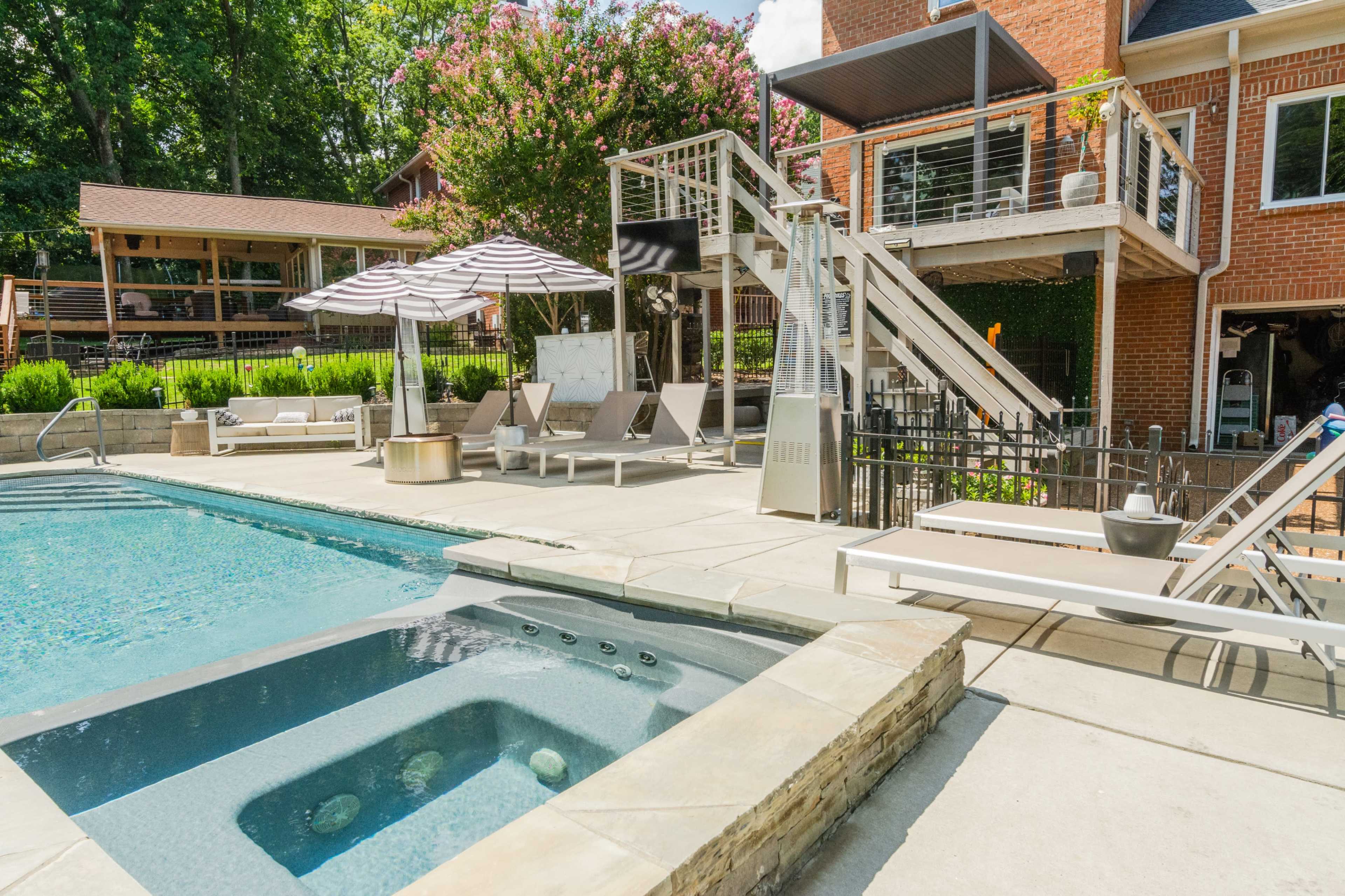 A backyard pool area with a hot tub, lounge chairs, and a patio, surrounded by greenery and a two-story deck.