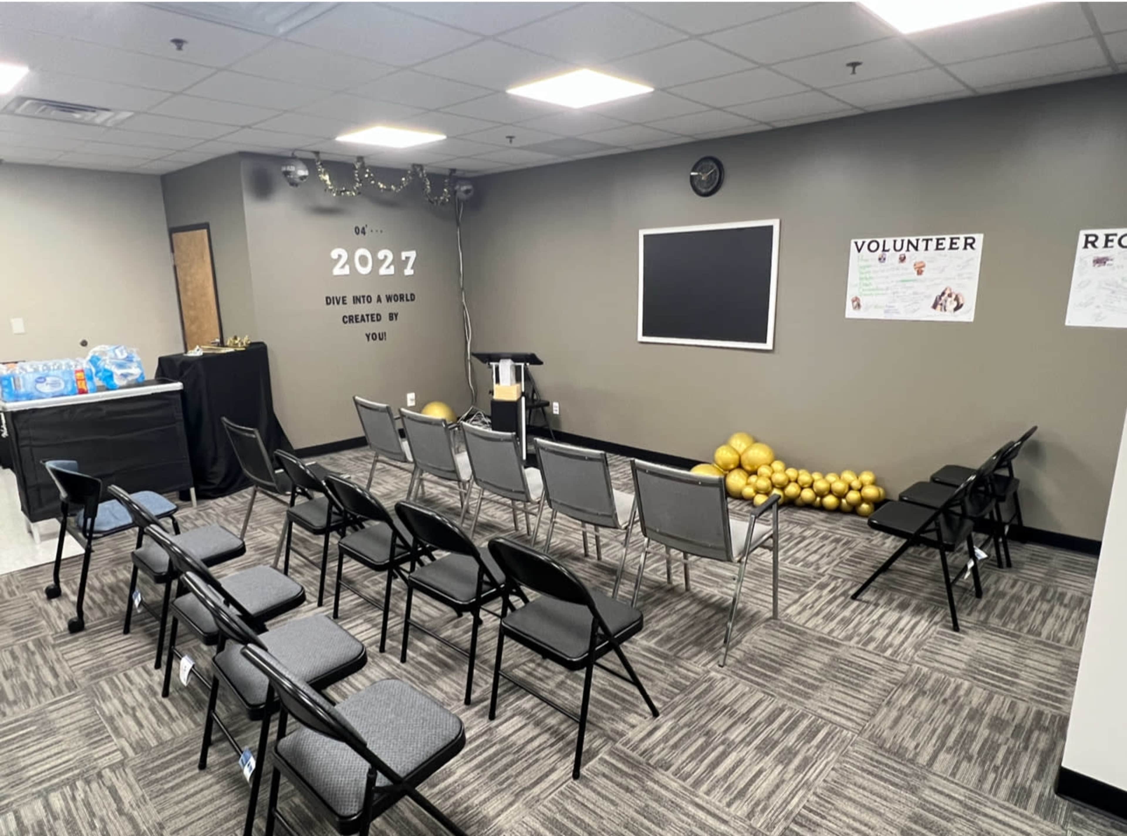 A conference room set up with several gray chairs arranged in rows, a black podium at the front, and a wall featuring decorative elements including a chalkboard and posters.