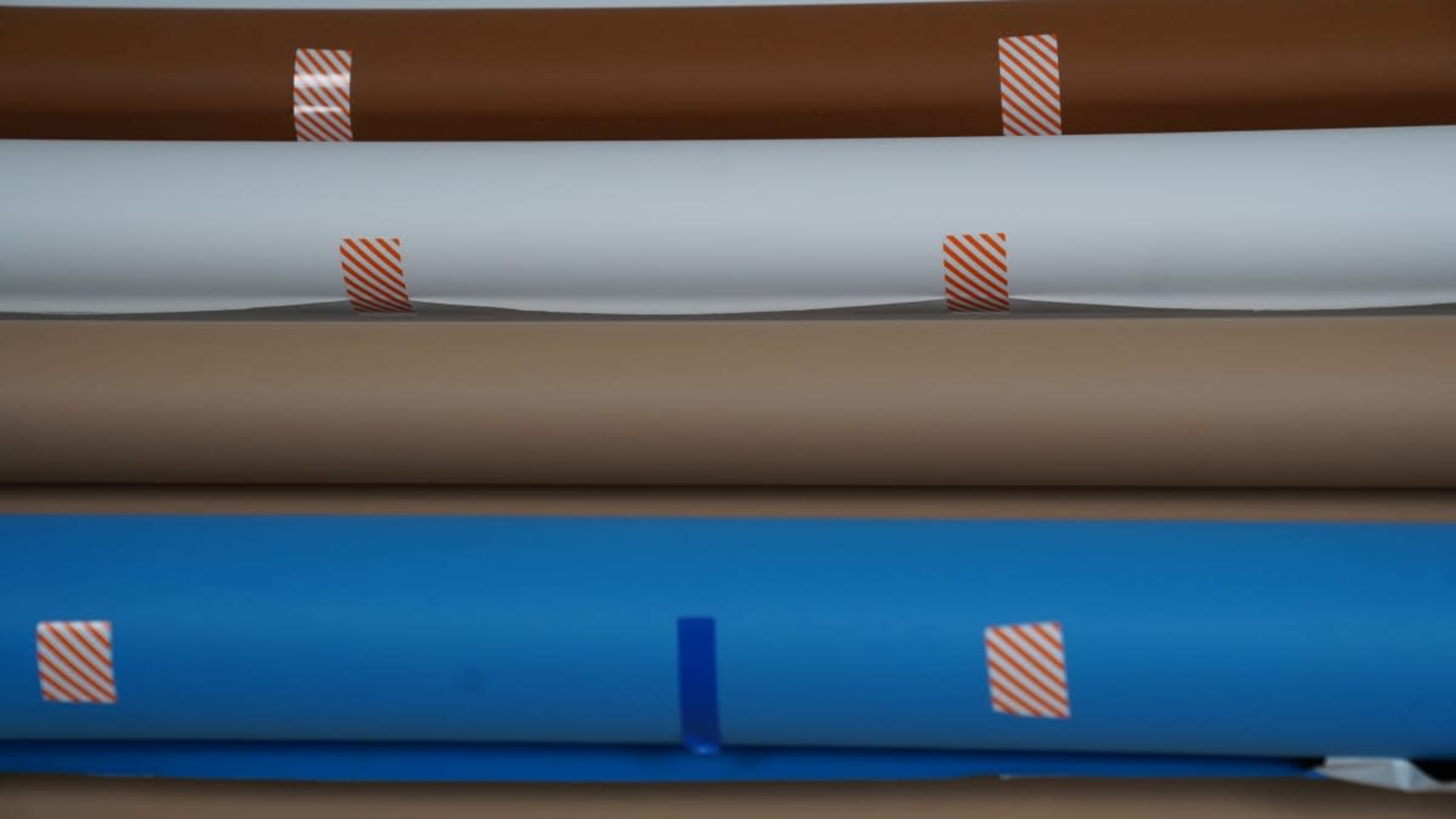 The image shows several rolls of colored paper stacked horizontally, including brown, white, and blue, secured with orange striped tape.