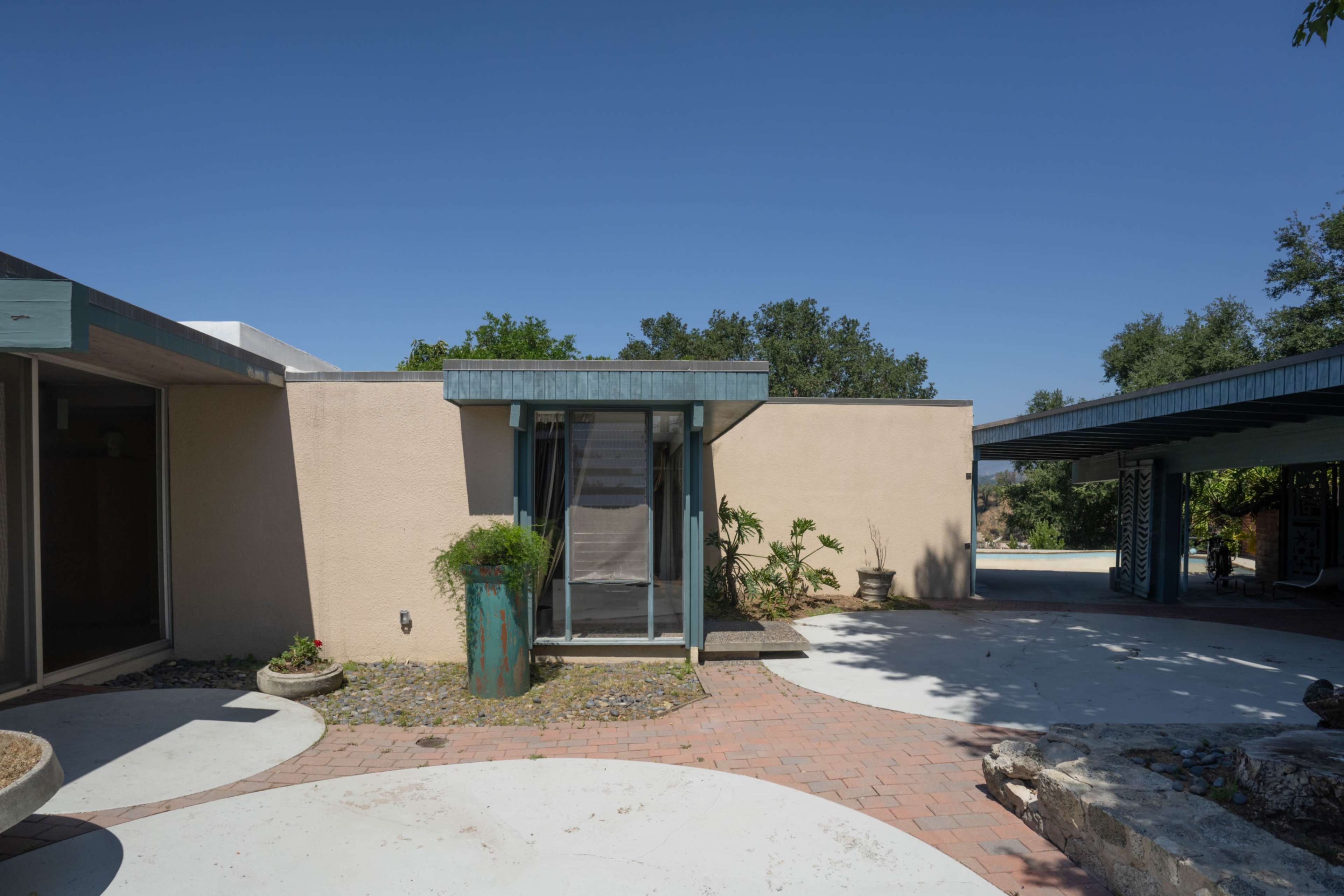 The image shows a modern architectural design featuring a light-colored exterior with a central entrance, surrounded by greenery and a paved outdoor area.