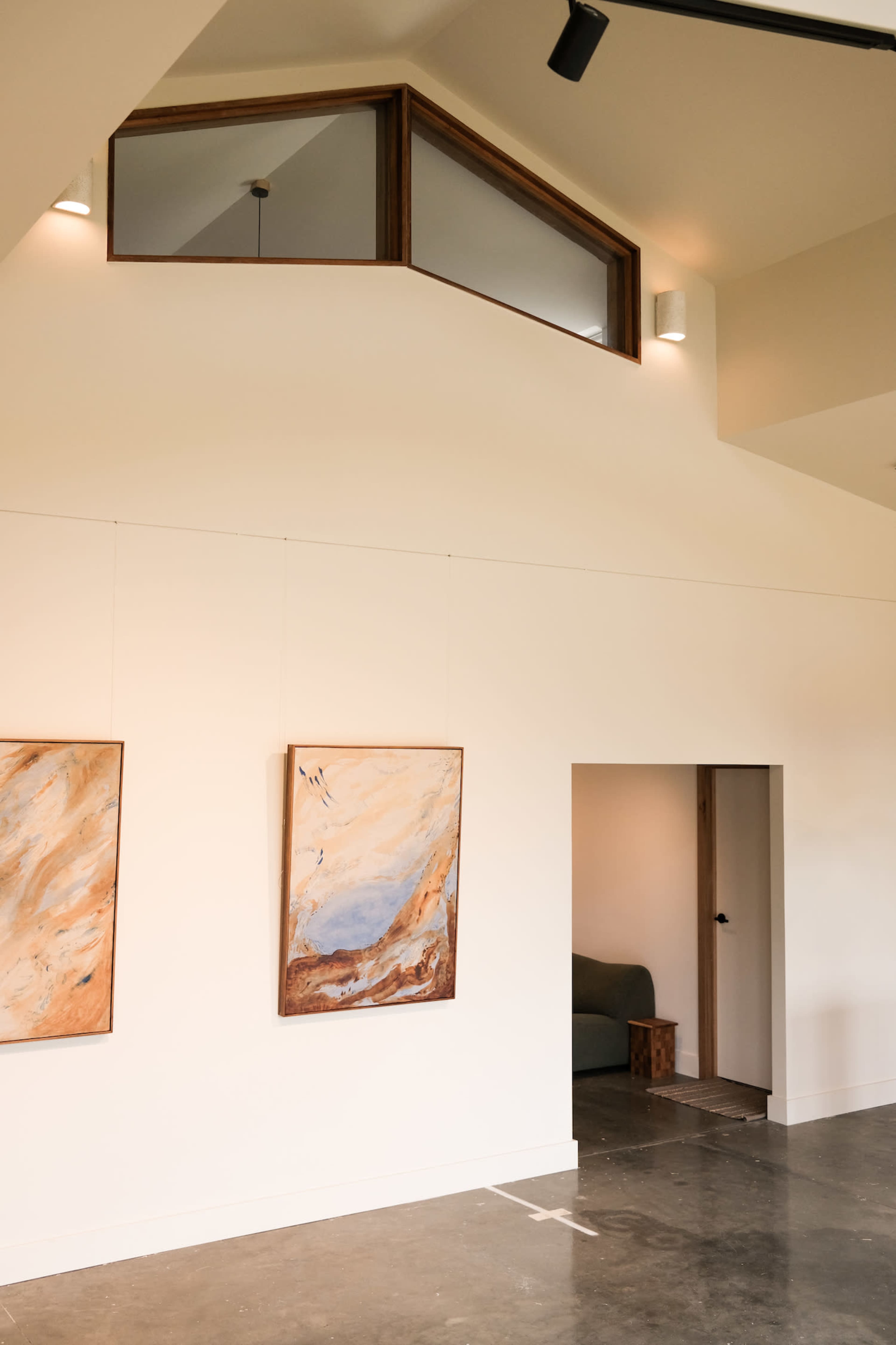 An interior space with a high ceiling, two abstract paintings on a white wall, and an open doorway leading to another area.