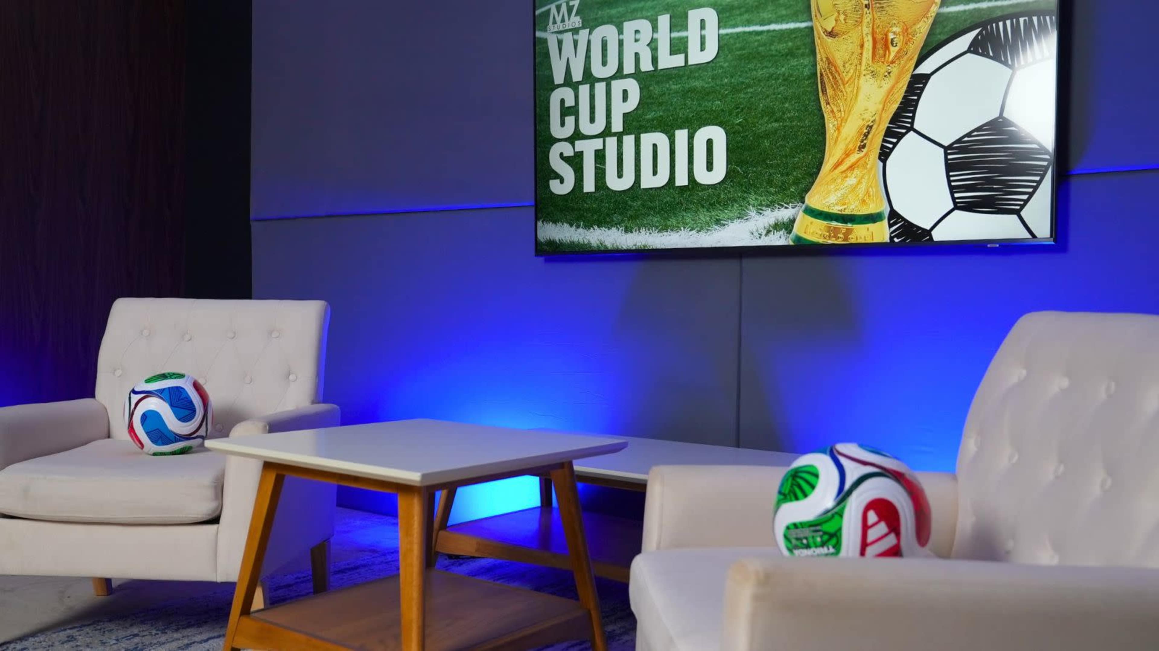 The image shows a television screen displaying "World Cup Studio" alongside a soccer trophy and two colorful soccer balls on a table in a modern seating area.